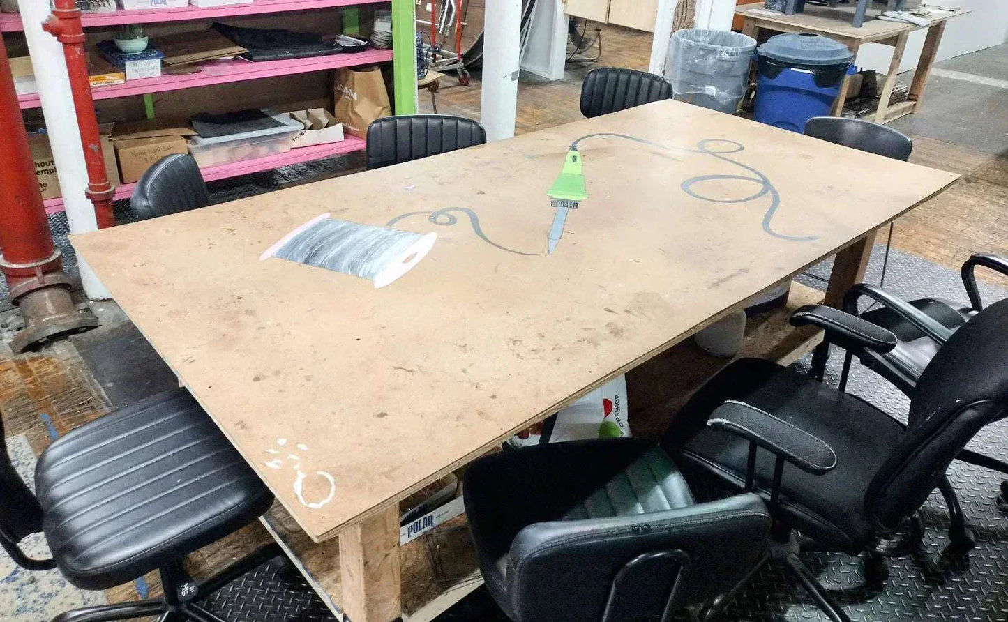 Wooden workshop table with painted tape, needle, and thread illustrations surrounded by black office chairs in an art studio setting.