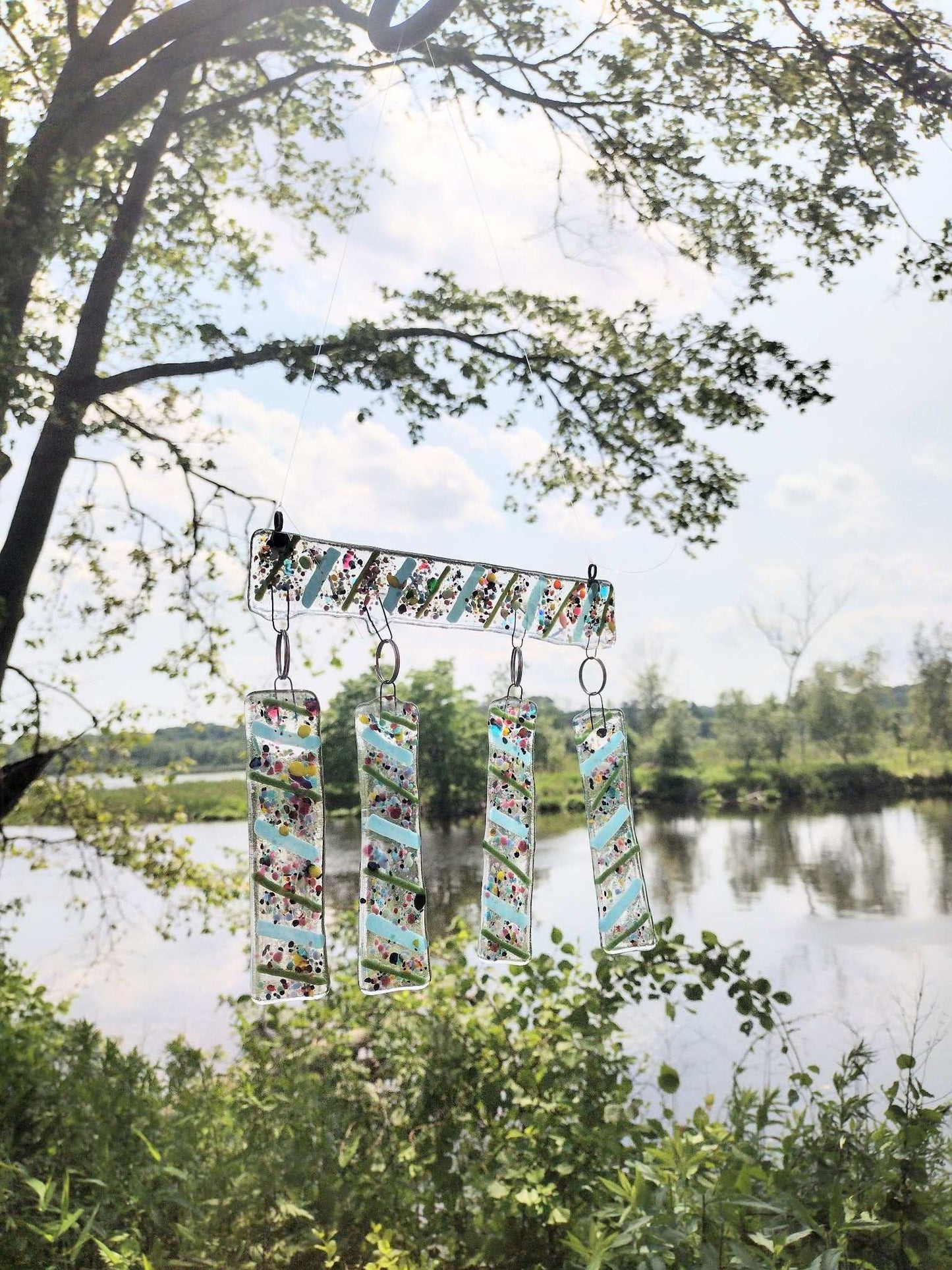 colorful fused glass windchimes hanging outdoors by a lake with trees in the background