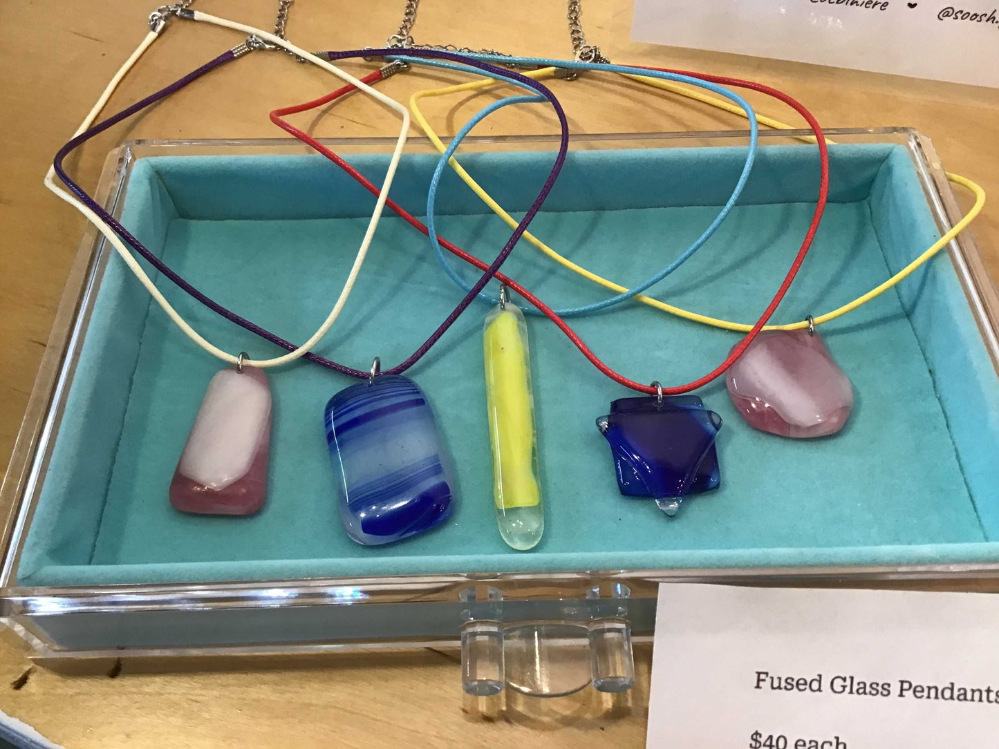 Colorful fused glass pendants on cords displayed in a blue tray with chains