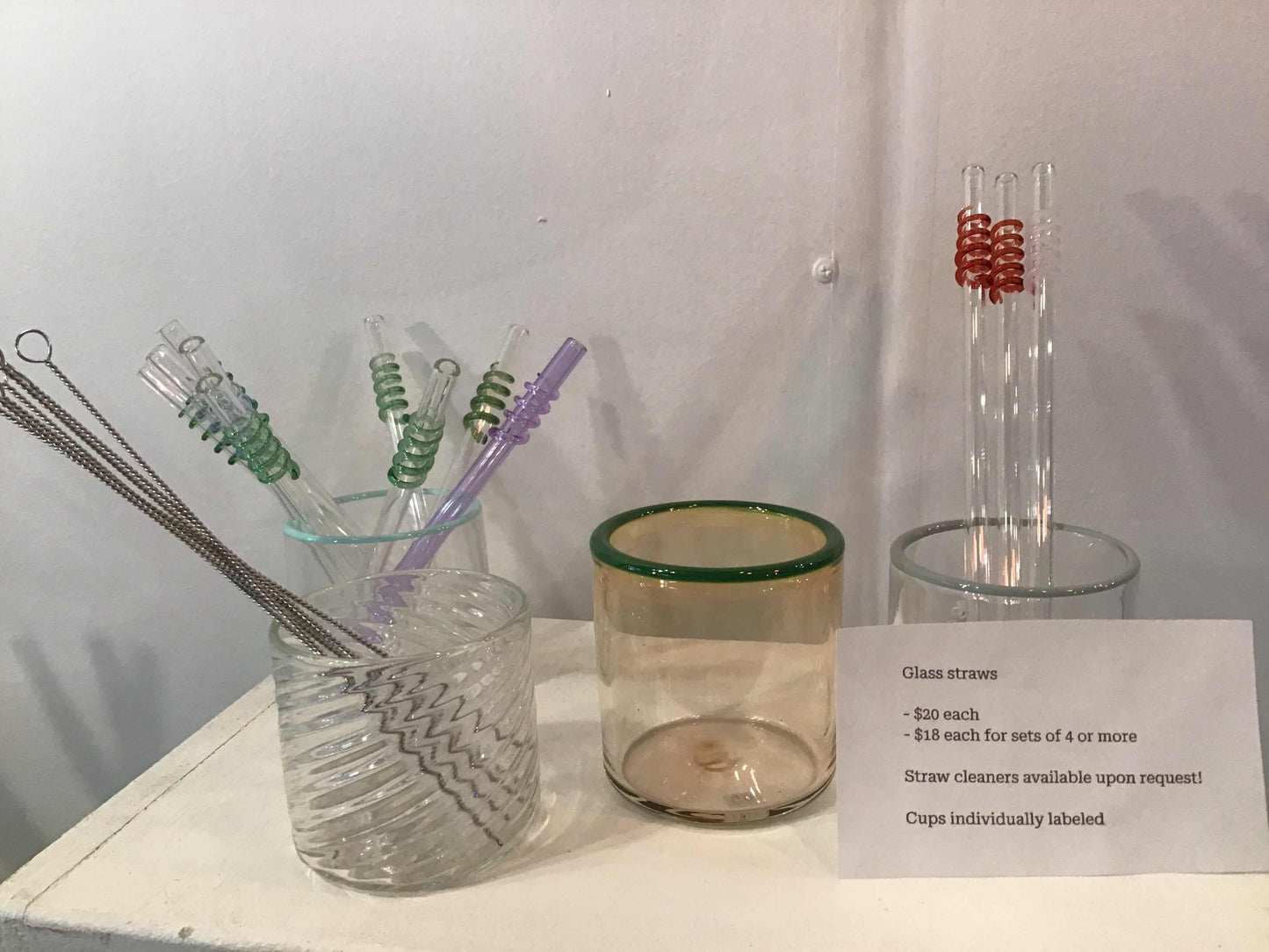 Glass reusable straws with coil decoration and cleaning brushes in clear holders on a table with pricing information.