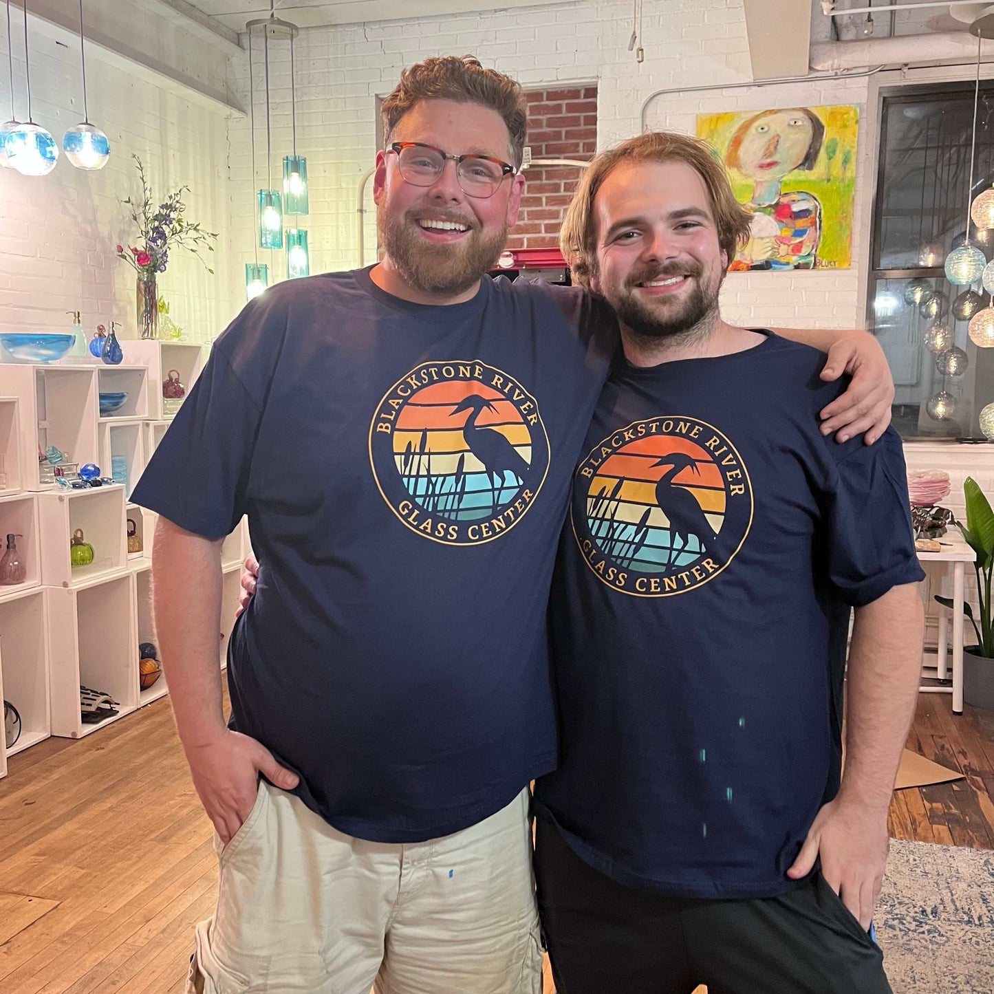 Two smiling men wearing Blackstone River Glass Center t-shirts standing together inside a glass blowing studio during a happy hour glass blowing event