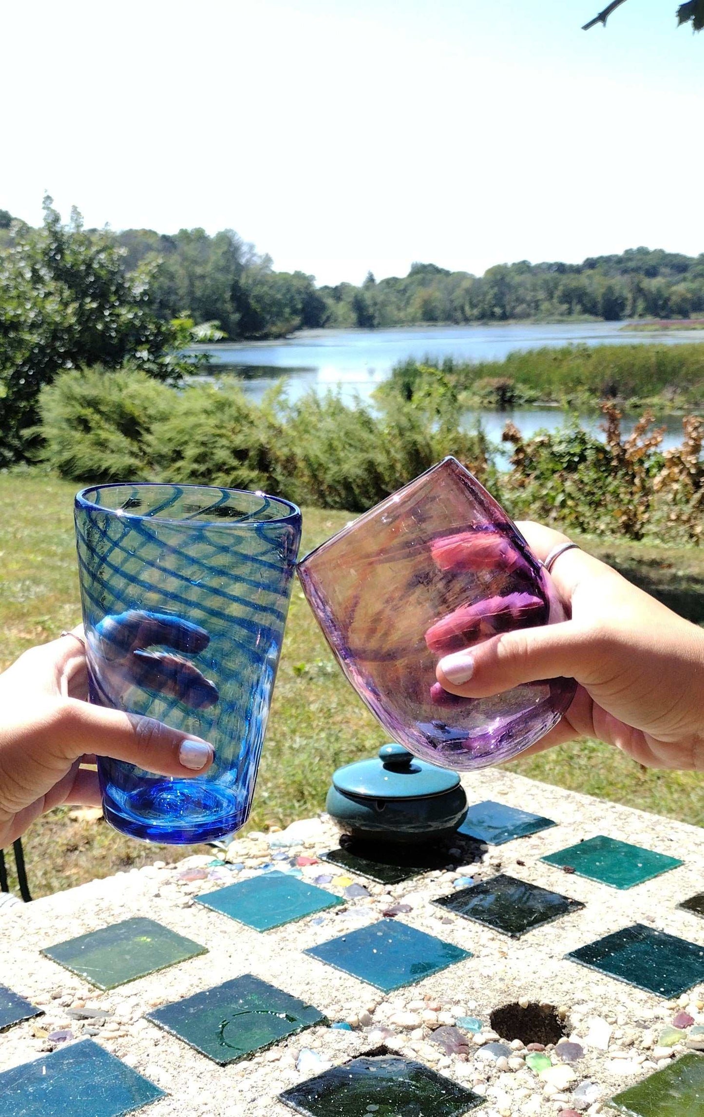 Couple clinking colorful hand-blown glasses outdoors by a lake for Date Night Glassblowing class