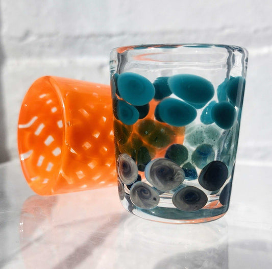 Clear flameworked shot glass with blue and gray decorative dots next to an orange textured glass