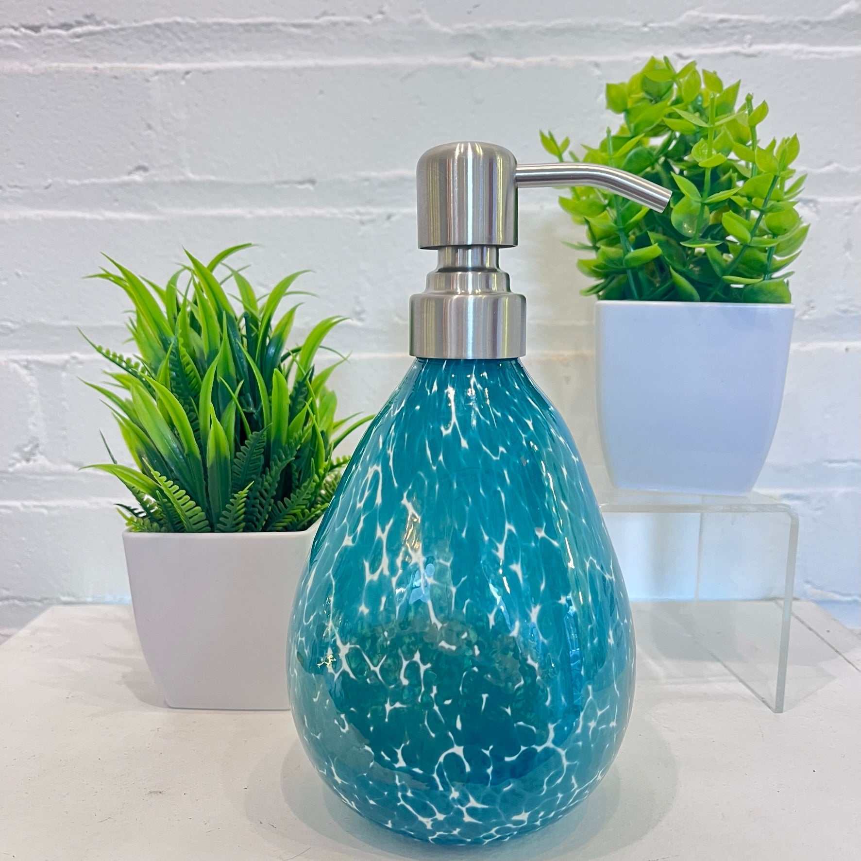 Hand blown glass teal speckled soap dispenser with stainless steel pump next to potted plants