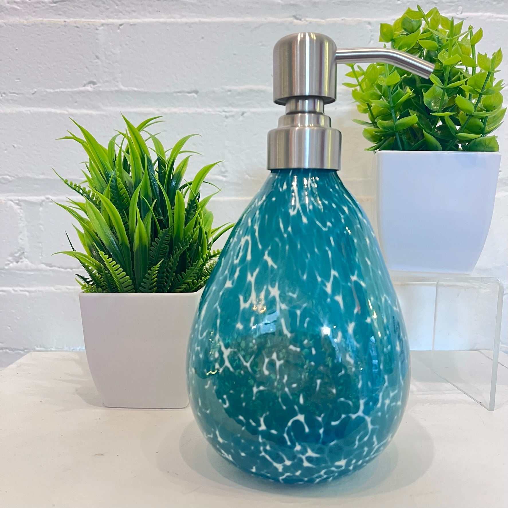 Hand blown glass teal speckled soap dispenser with stainless steel pump and decorative plants background