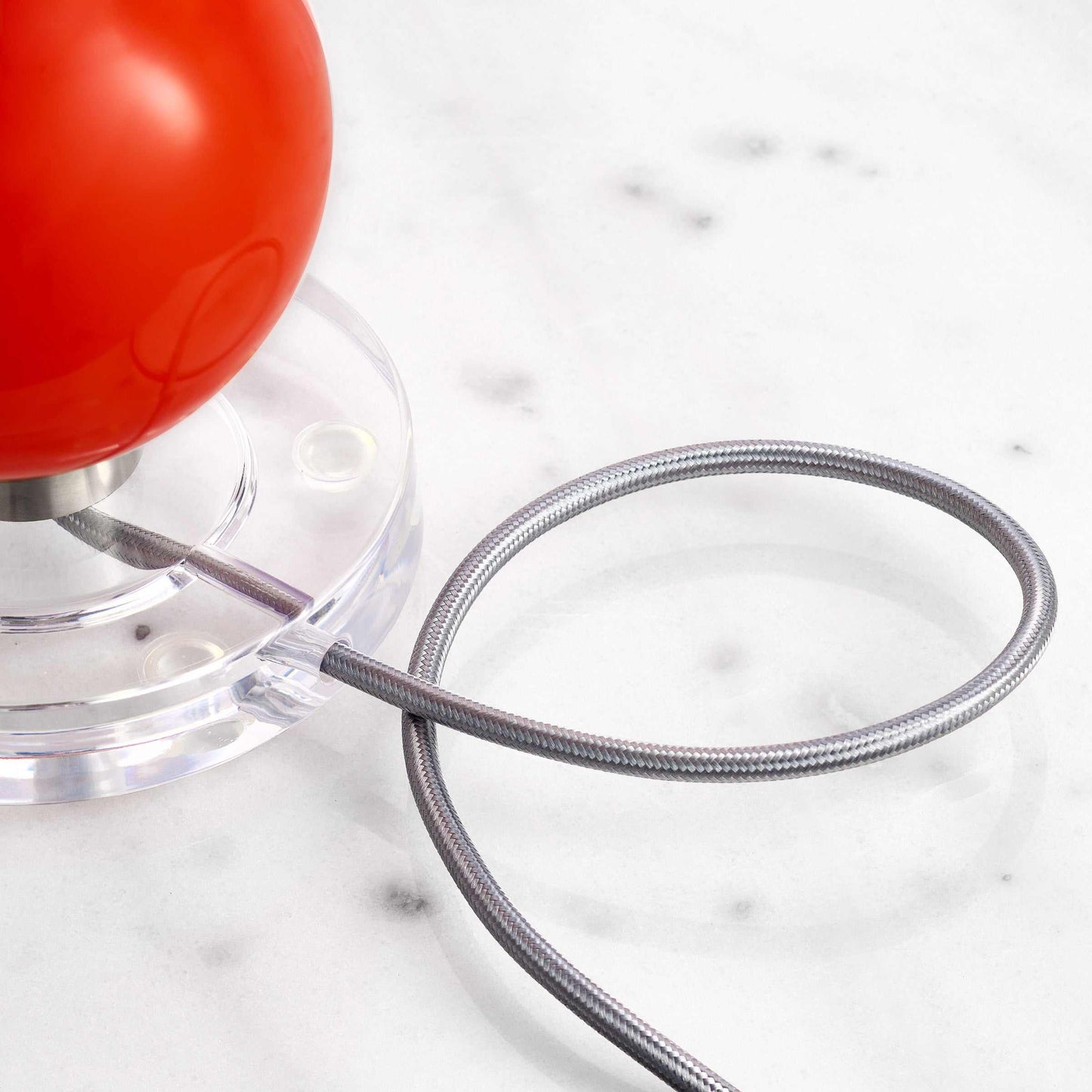 Close-up of Carmen Table Lamp's clear acrylic base with vibrant red glass globe and gray textile cord on white marble surface
