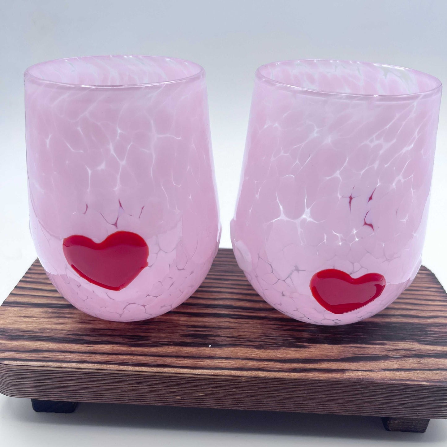 Pair of hand-blown pink stemless wine glasses with red heart accents on wooden stand