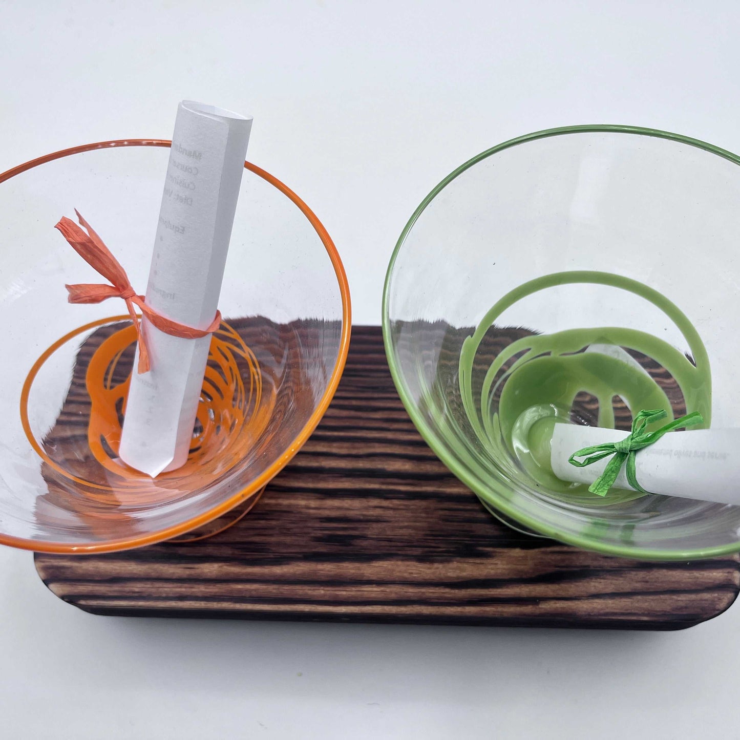 Pair of hand blown stemless martini glasses in orange and green with rolled notes, displayed on a wooden tray