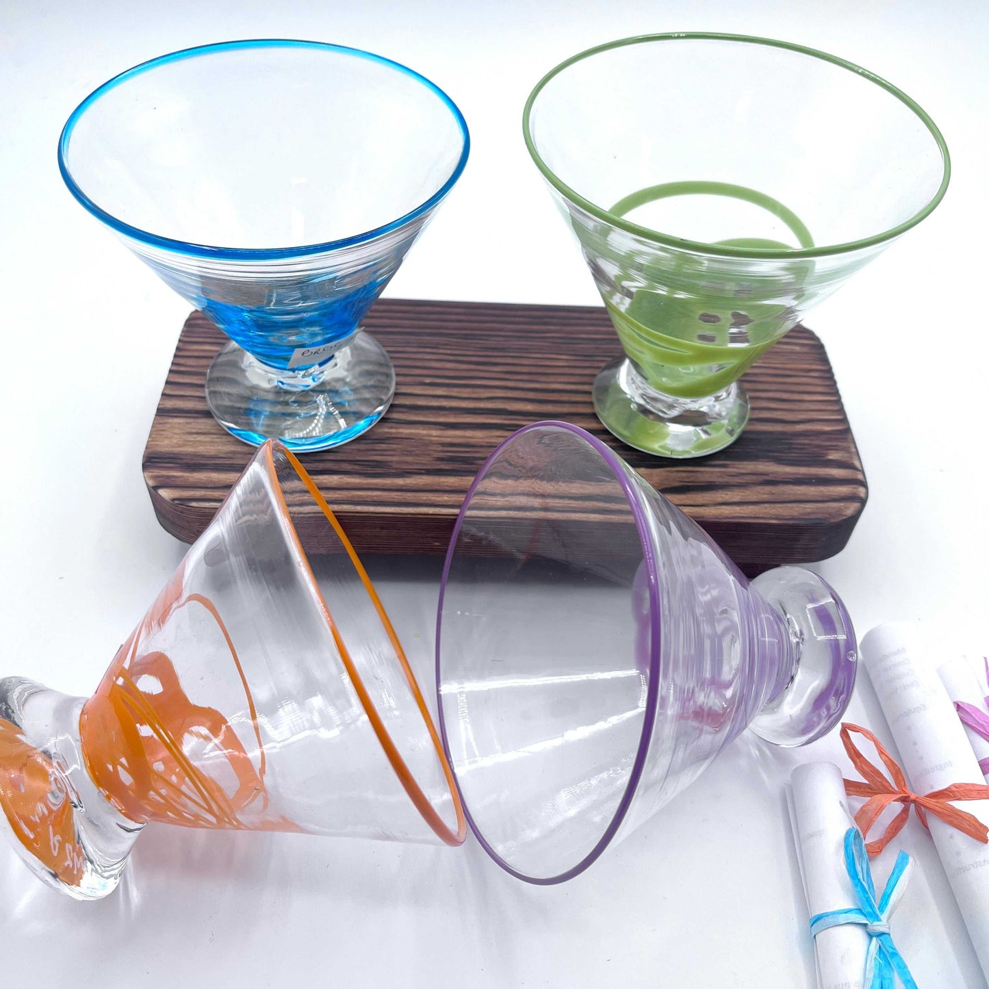 Set of hand blown stemless martini glasses with colored rims on wooden board and white surface.