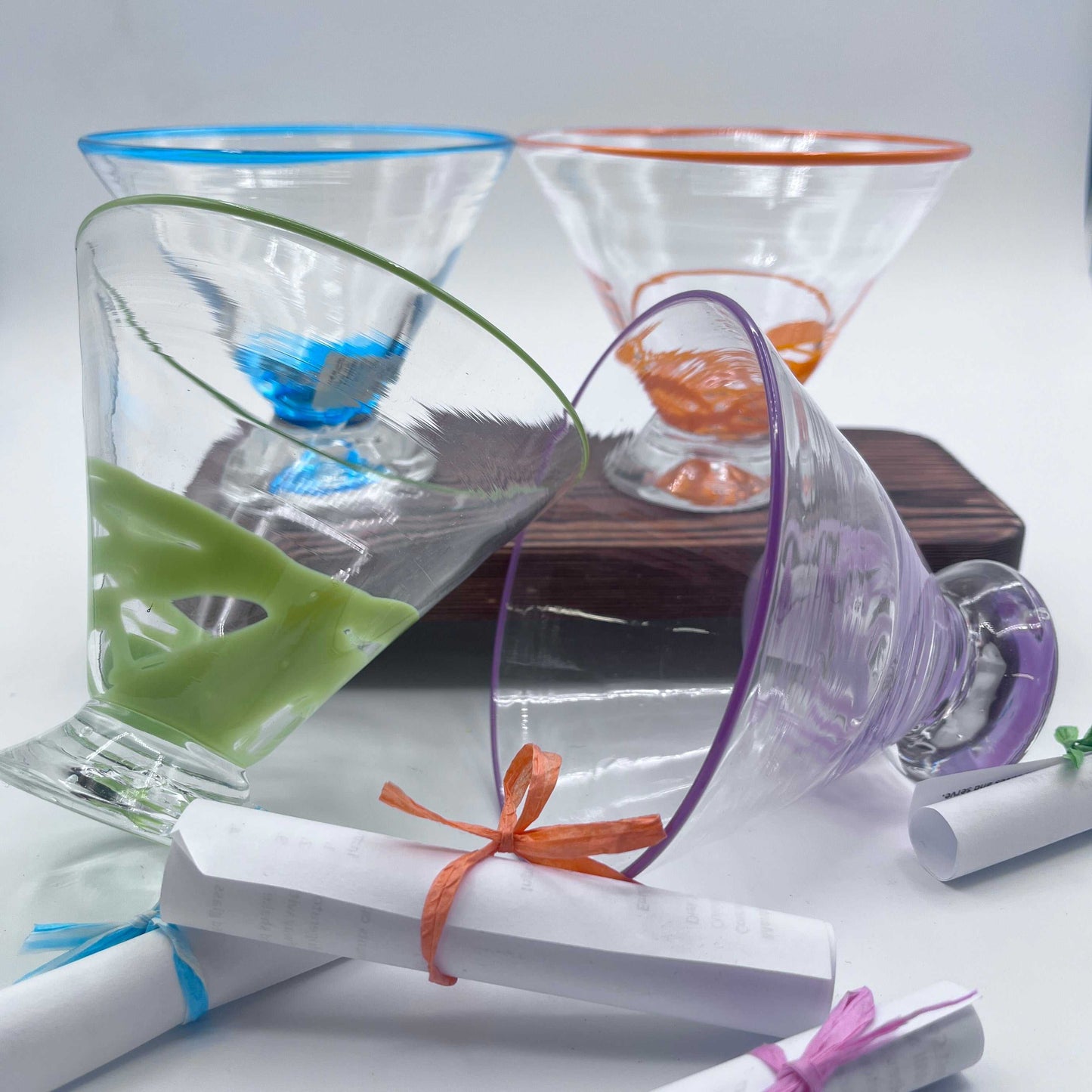 Set of colorful hand blown stemless martini glasses with decorative rims, displayed with tied scrolls on a white background