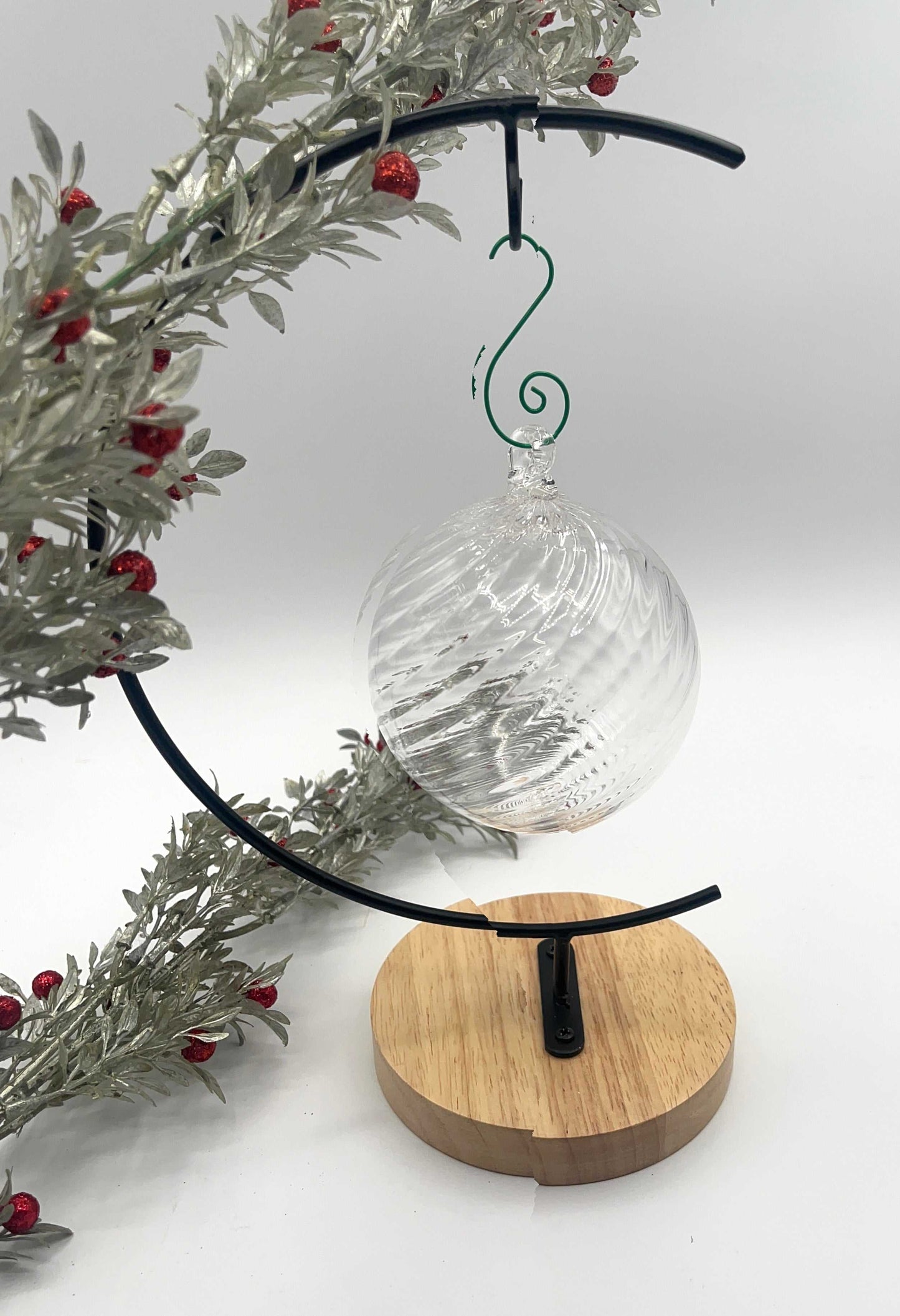 Blown glass ornament with clear spiral twist hanging on black metal stand with wooden base and decorative foliage with red berries.