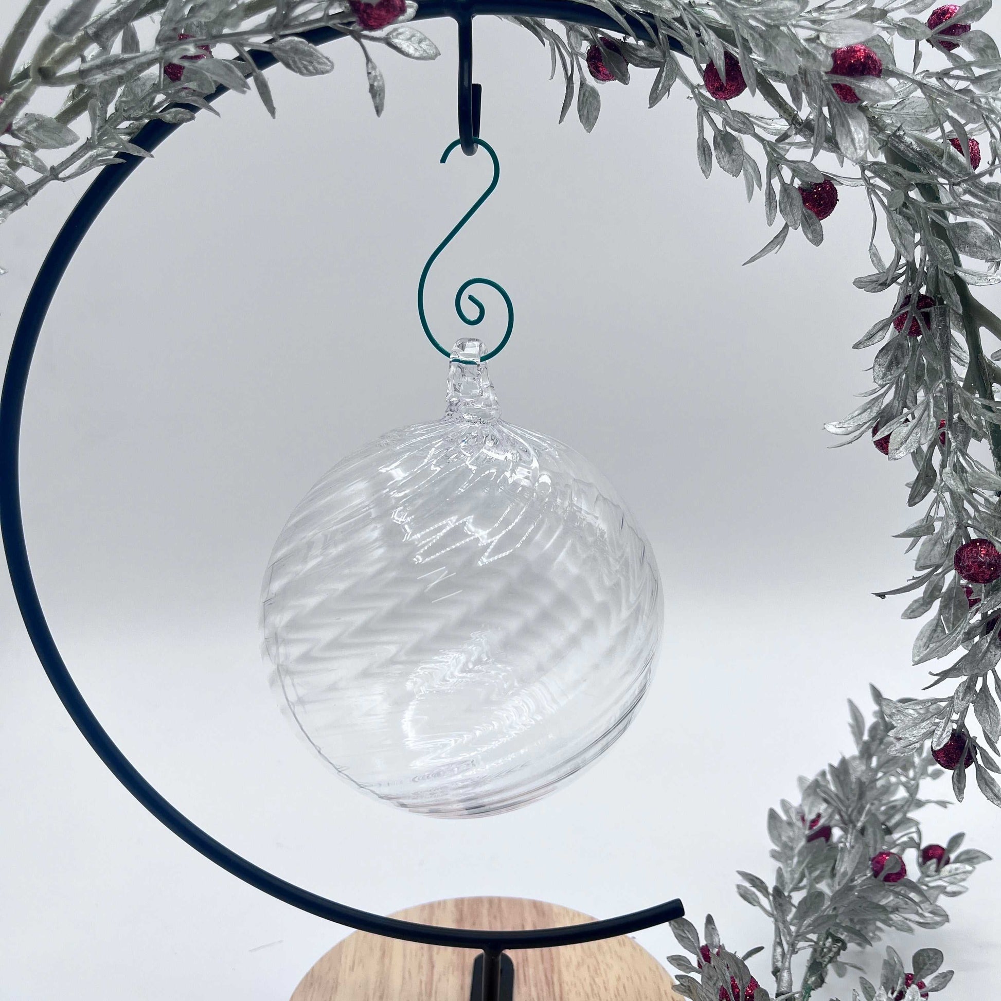Clear spiral twist blown glass ornament hanging within decorative silver and red berry wreath on black circular stand