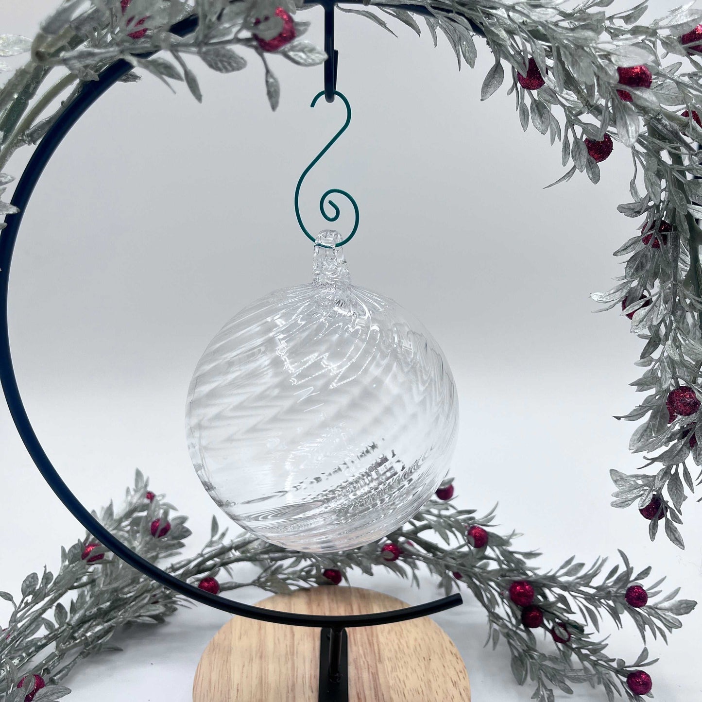 Clear blown glass ornament with spiral twist design hanging on black metal stand with festive silver and red berry wreath background