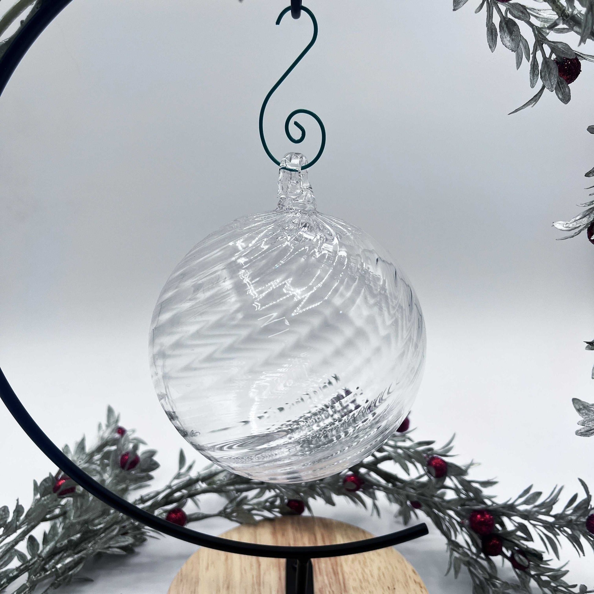 Clear blown glass ornament with spiral twist design hanging on black stand with festive greenery background