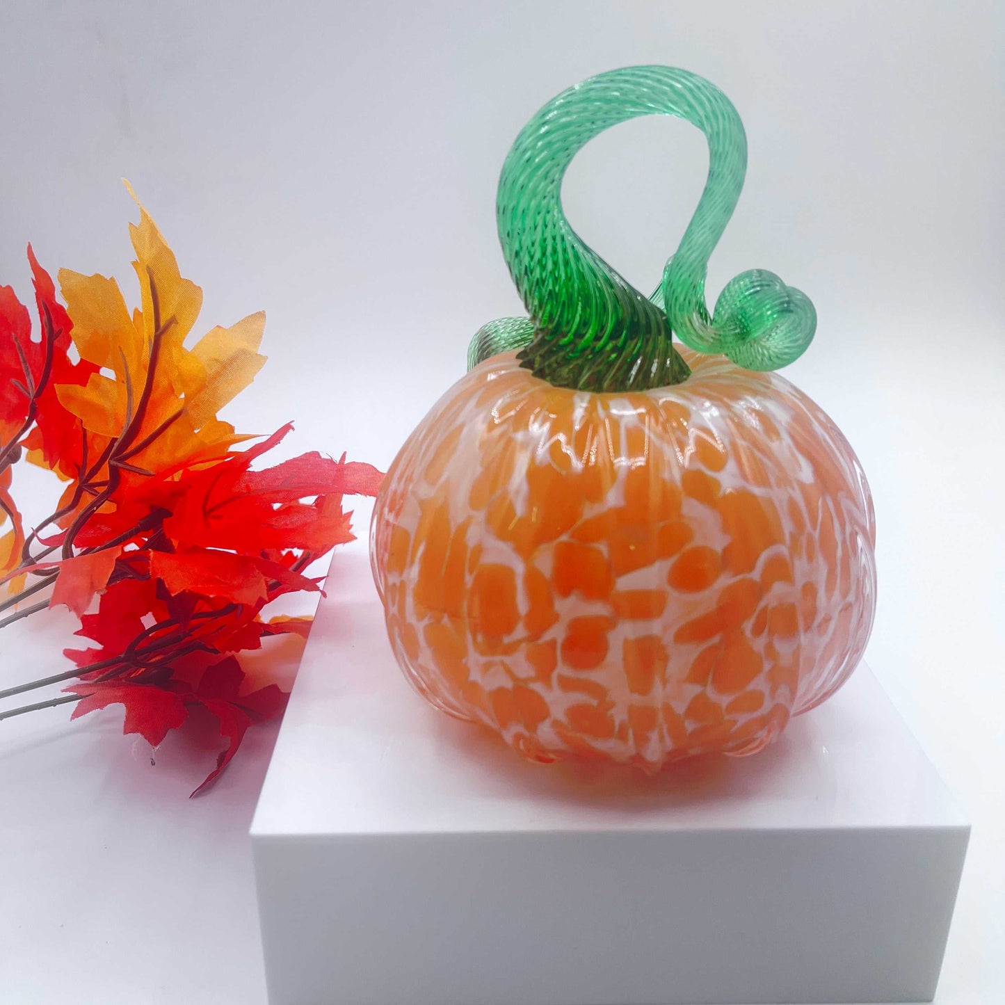 Hand-blown art glass pumpkin with orange cream over white color and green stem, displayed with autumn leaves on white surface