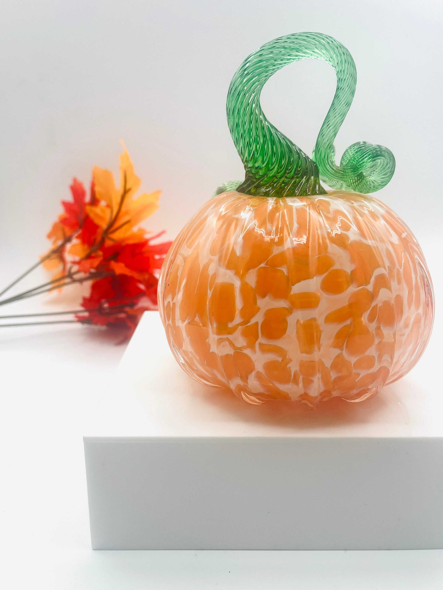 Hand-blown art glass pumpkin in orange creamsicle color with green twisted stem and autumn leaves background