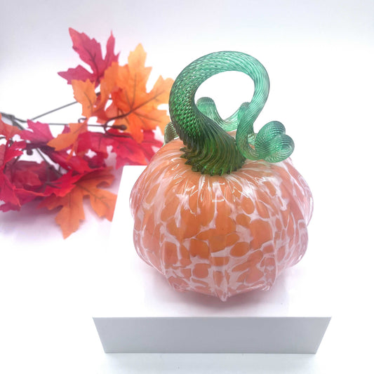 Hand-blown art glass pumpkin in orange creamsicle color with green stem and autumn leaves background