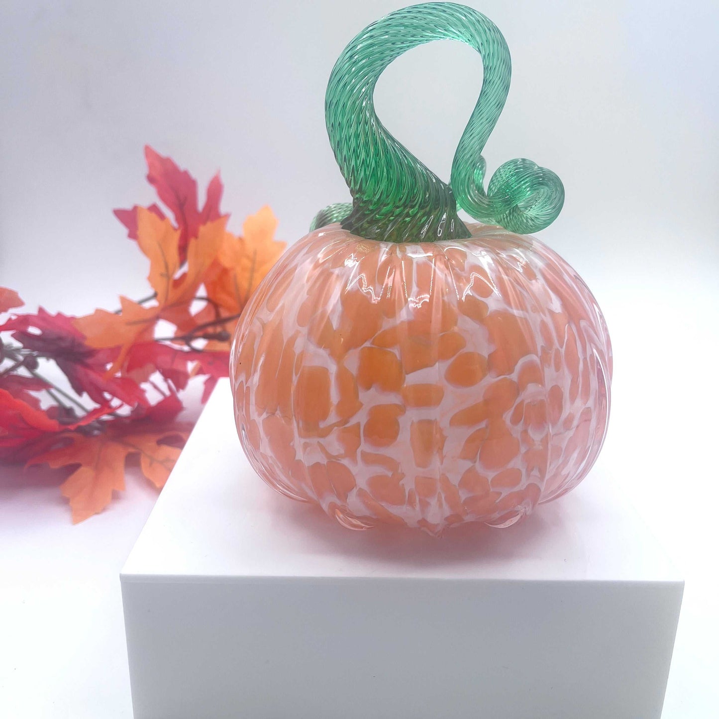 Hand-blown art glass pumpkin in orange creamsicle color with green twisted stem, autumn leaves background