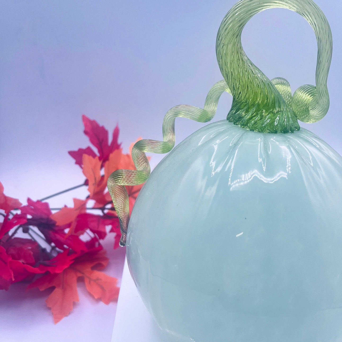 Large hand-blown glass pumpkin in celedon color with lime green stem and curled vine, autumn decor with red leaves background