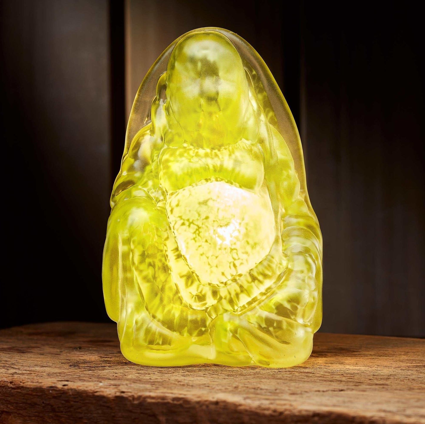 Olive hand blown glass Buddha light with smooth curves and warm glowing center on wooden surface.