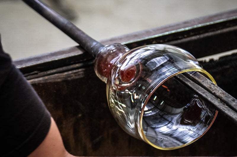 Close-up of a hand blowing glass during a Happy Hour Glass Blowing session with molten glass on a blowpipe in a workshop setting.
