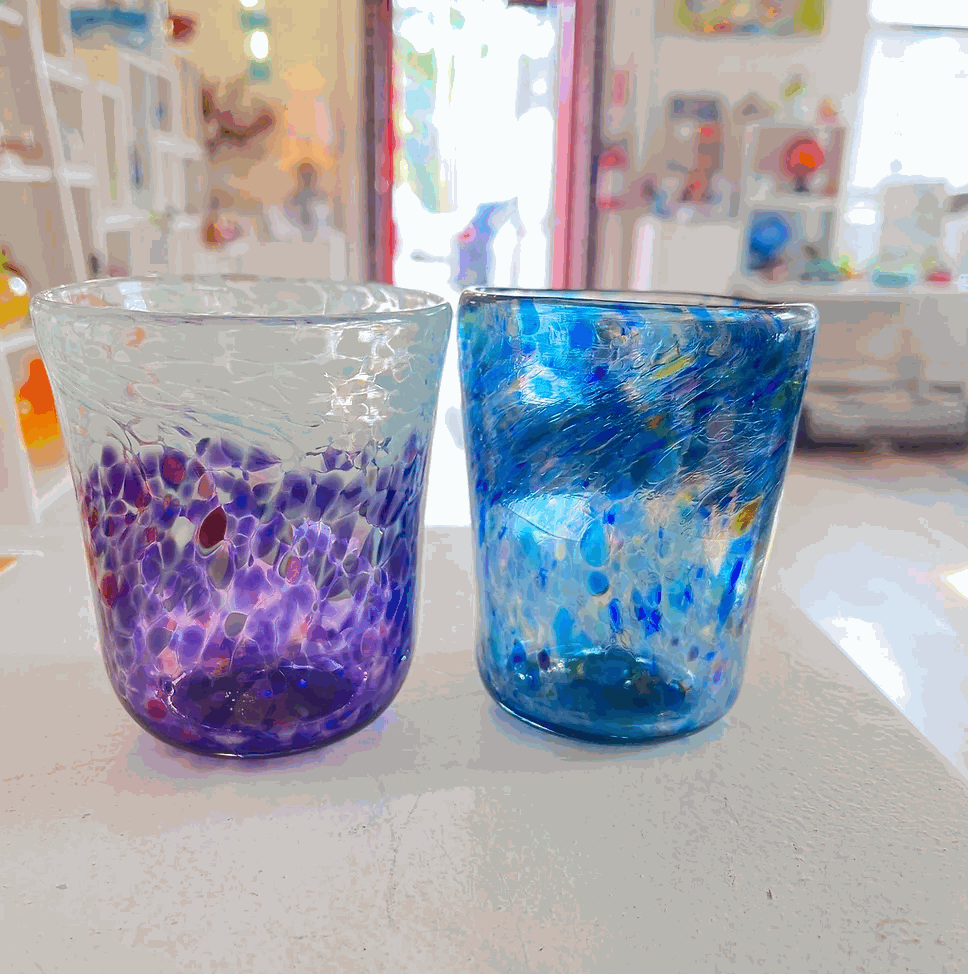 Two colorful handmade drinking glasses, one purple and one blue, displayed on a white surface in a craft studio setting.