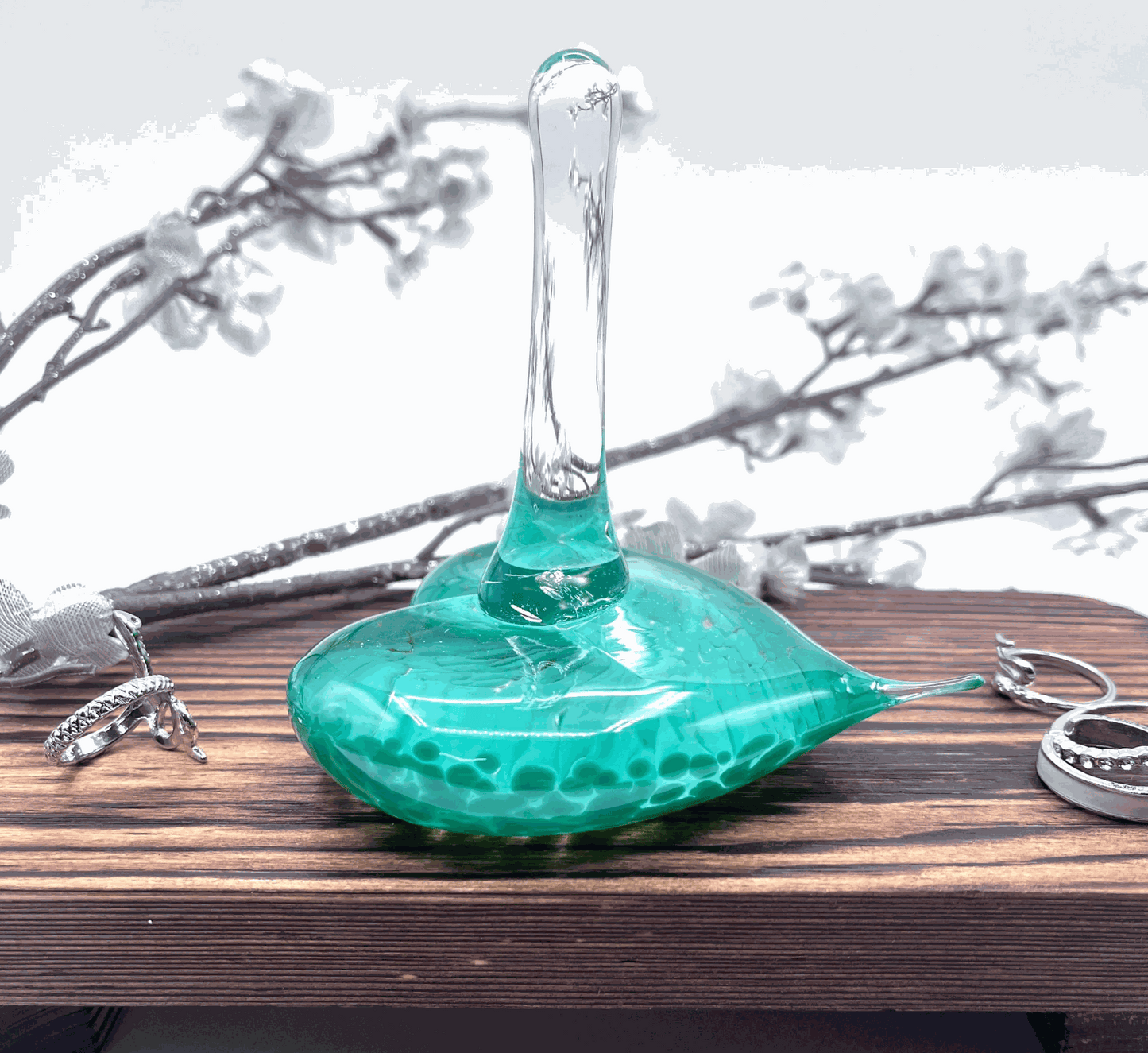 Jade green handmade glass heart ring stand with swirled design on wooden surface with rings and floral background
