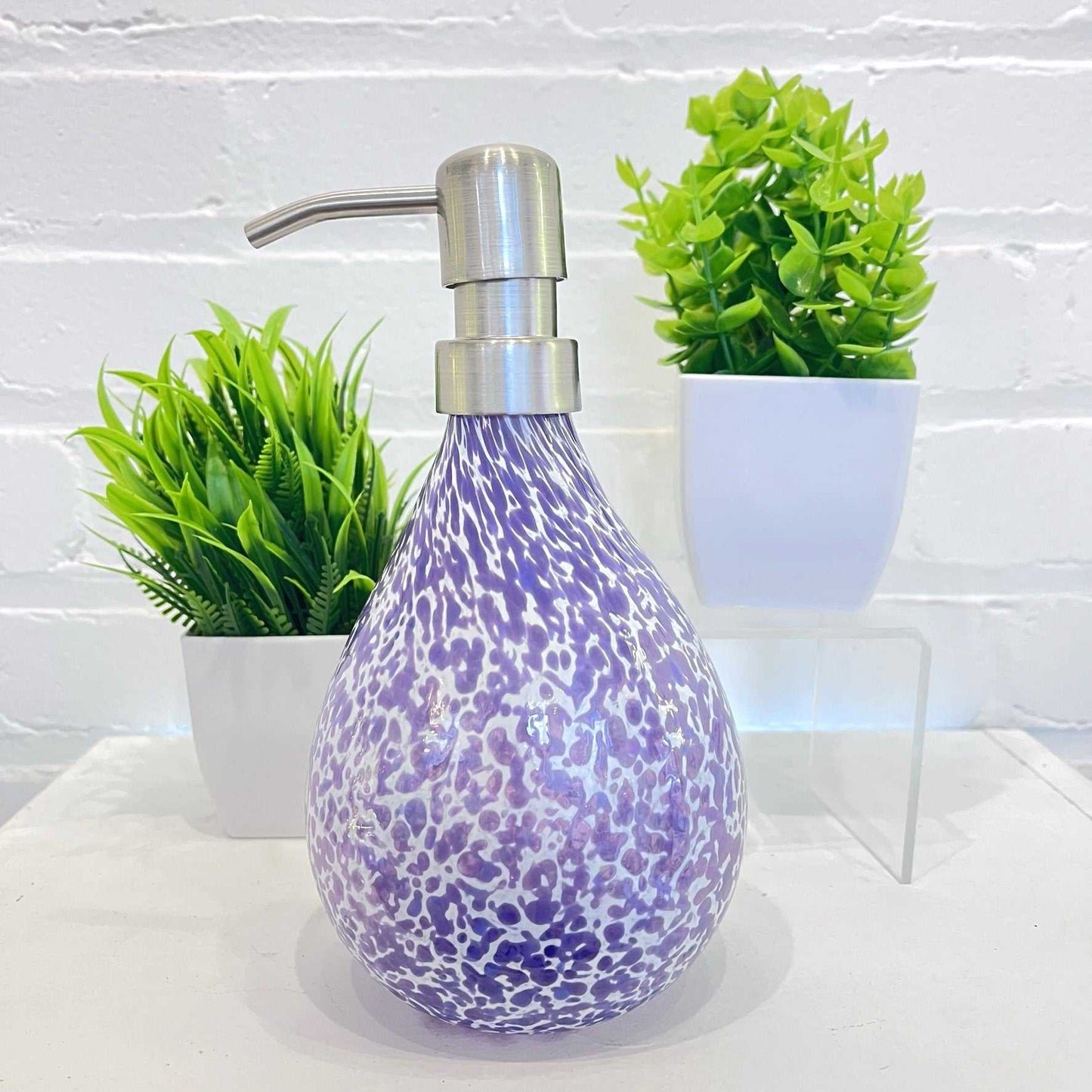 Hyacinth Riverstone hand blown glass soap dispenser with marbled purple and white pattern and stainless steel pump next to green potted plants
