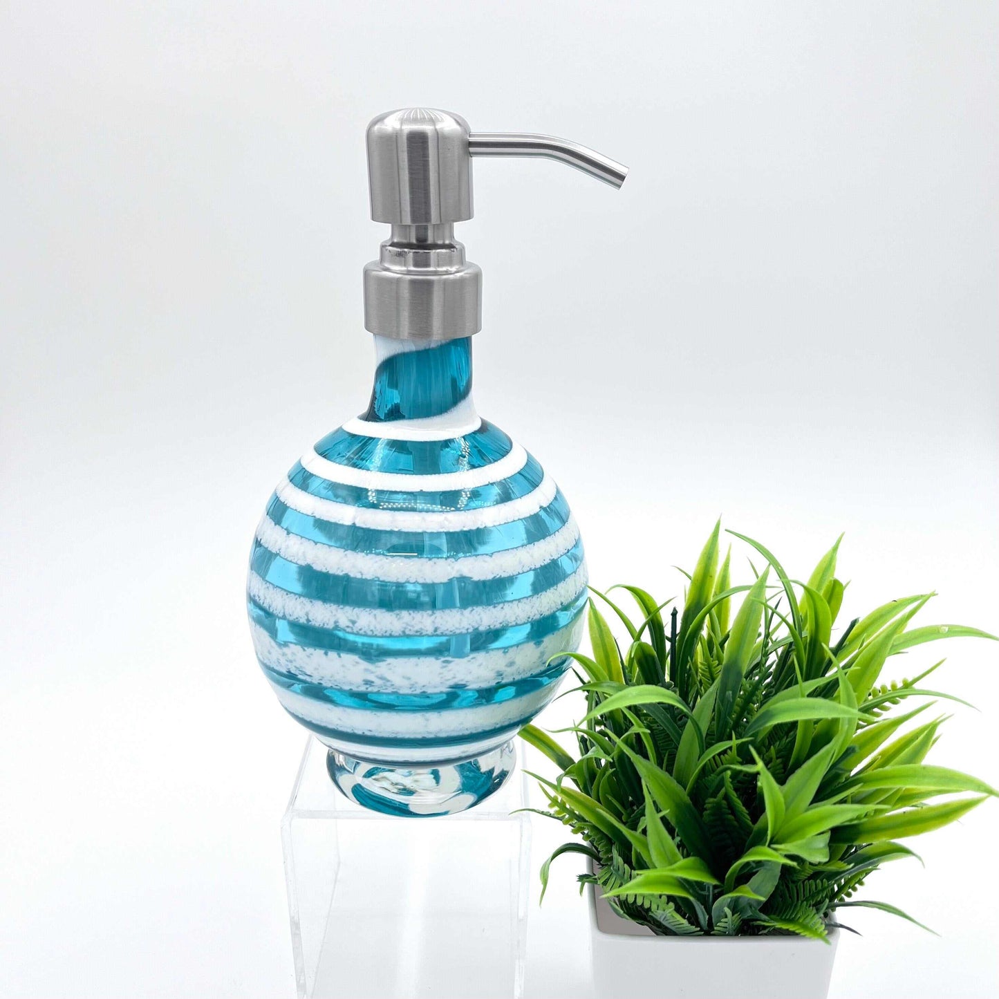 Boho handblown teal blue glass soap dispenser with distressed white spiral and stainless steel pump beside green plant