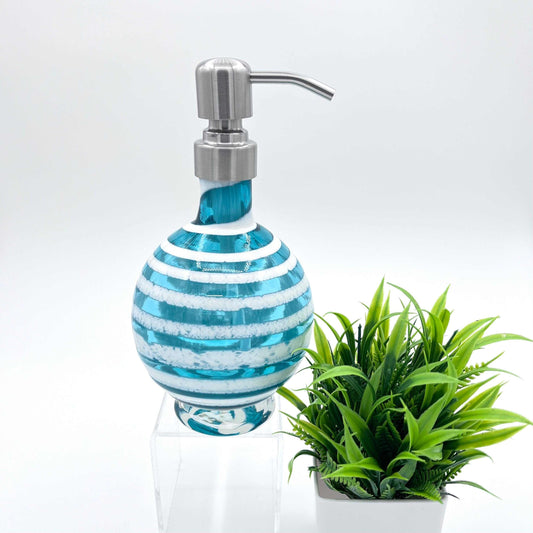 Boho handblown teal blue glass soap dispenser with distressed white spiral and stainless steel pump beside green plant