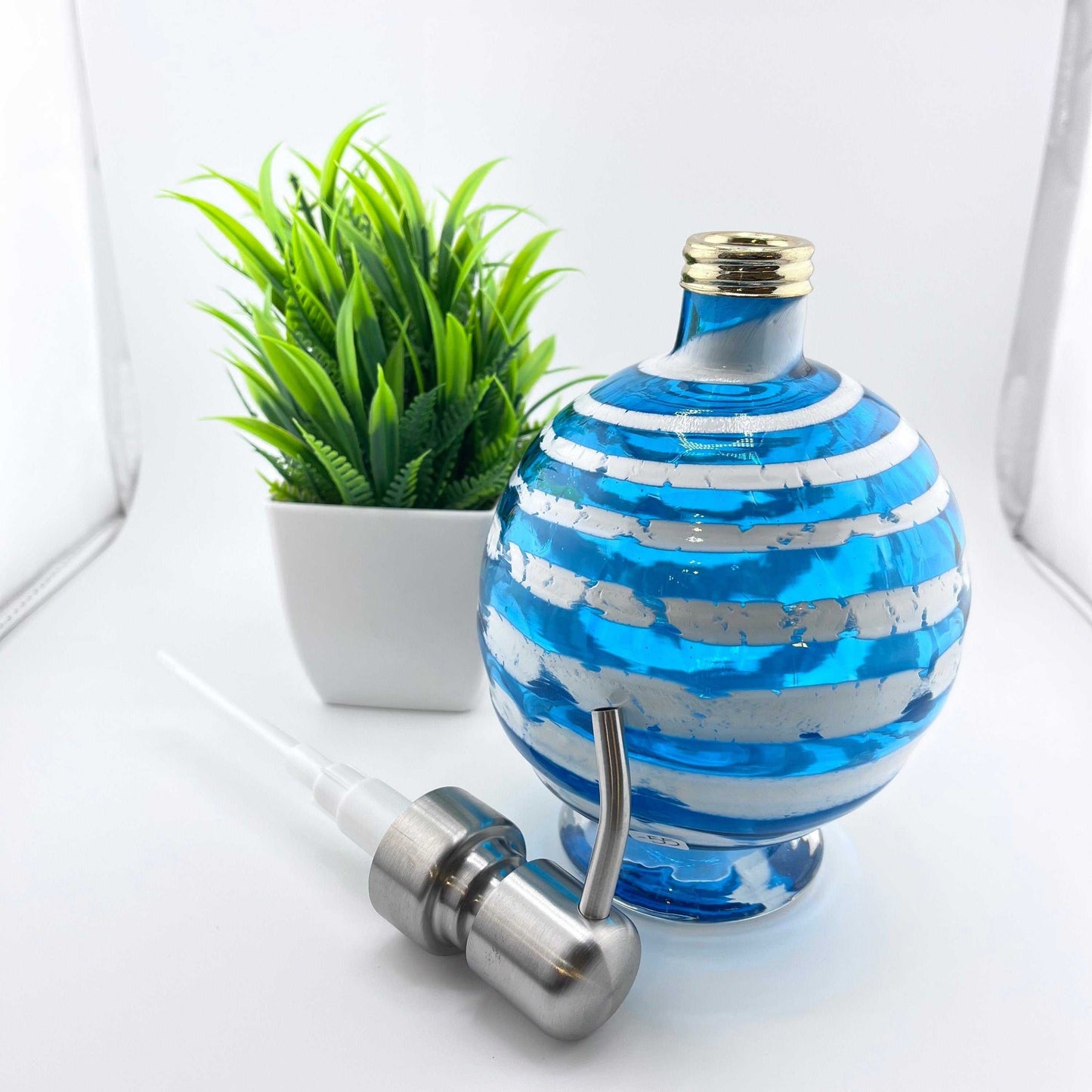 Boho handblown glass soap dispenser in aqua blue with distressed white spiral and removable metal pump next to a small green plant