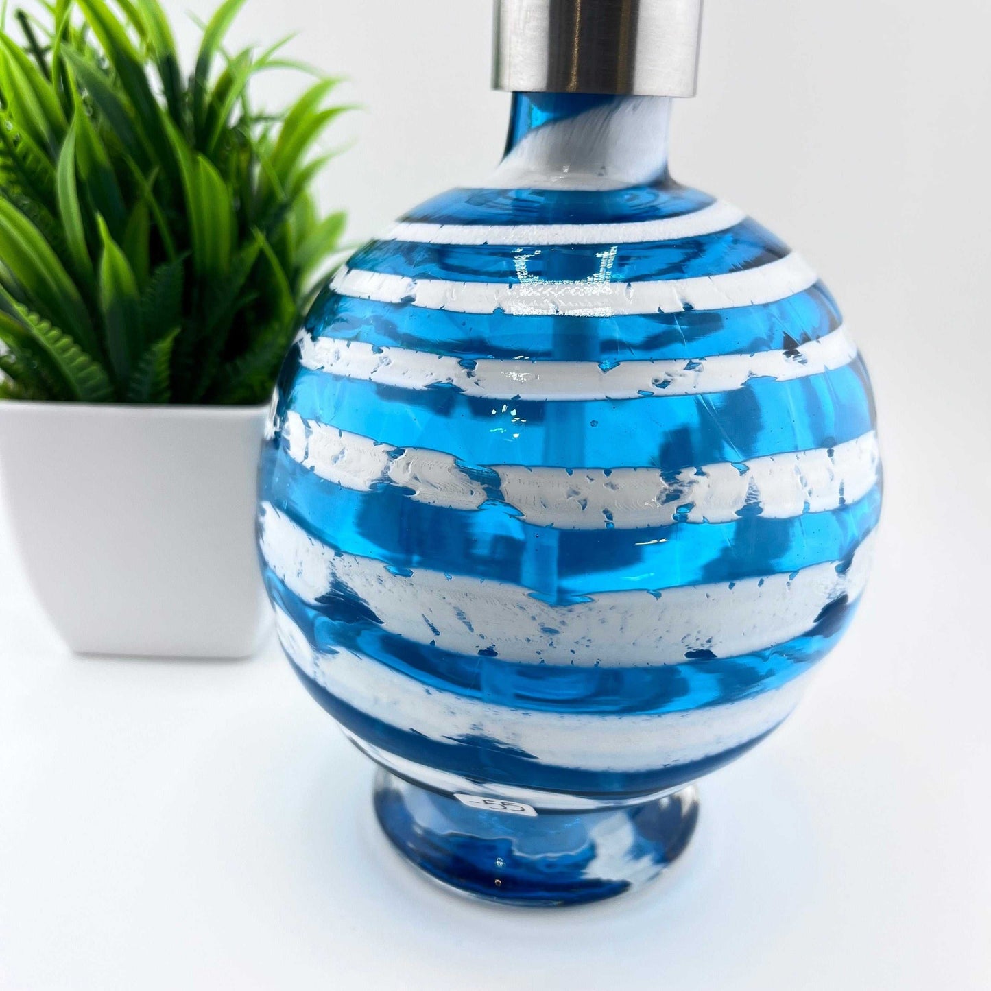 Boho handblown glass soap dispenser in aqua blue with distressed white spiral and stainless steel pump top next to green plant