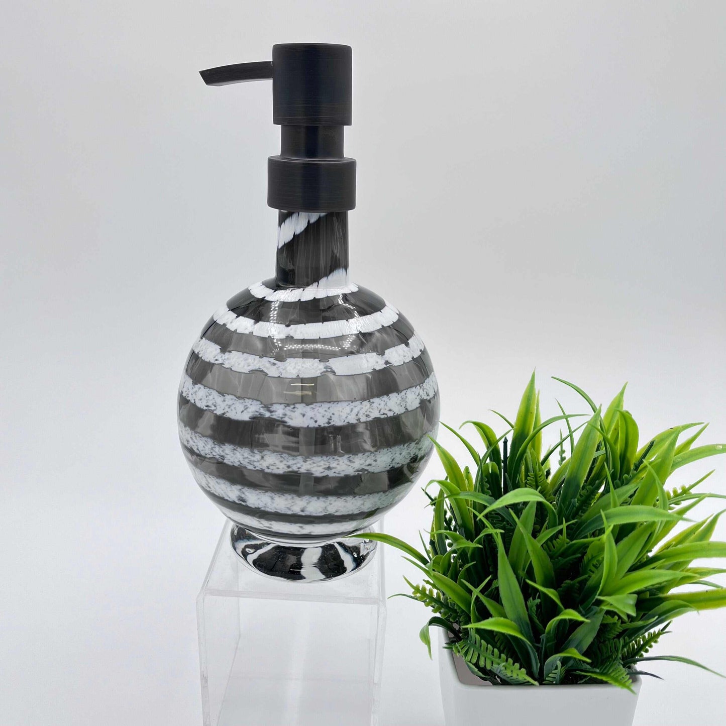 Boho handblown glass soap dispenser in gray with distressed white spiral and black pump next to green plant
