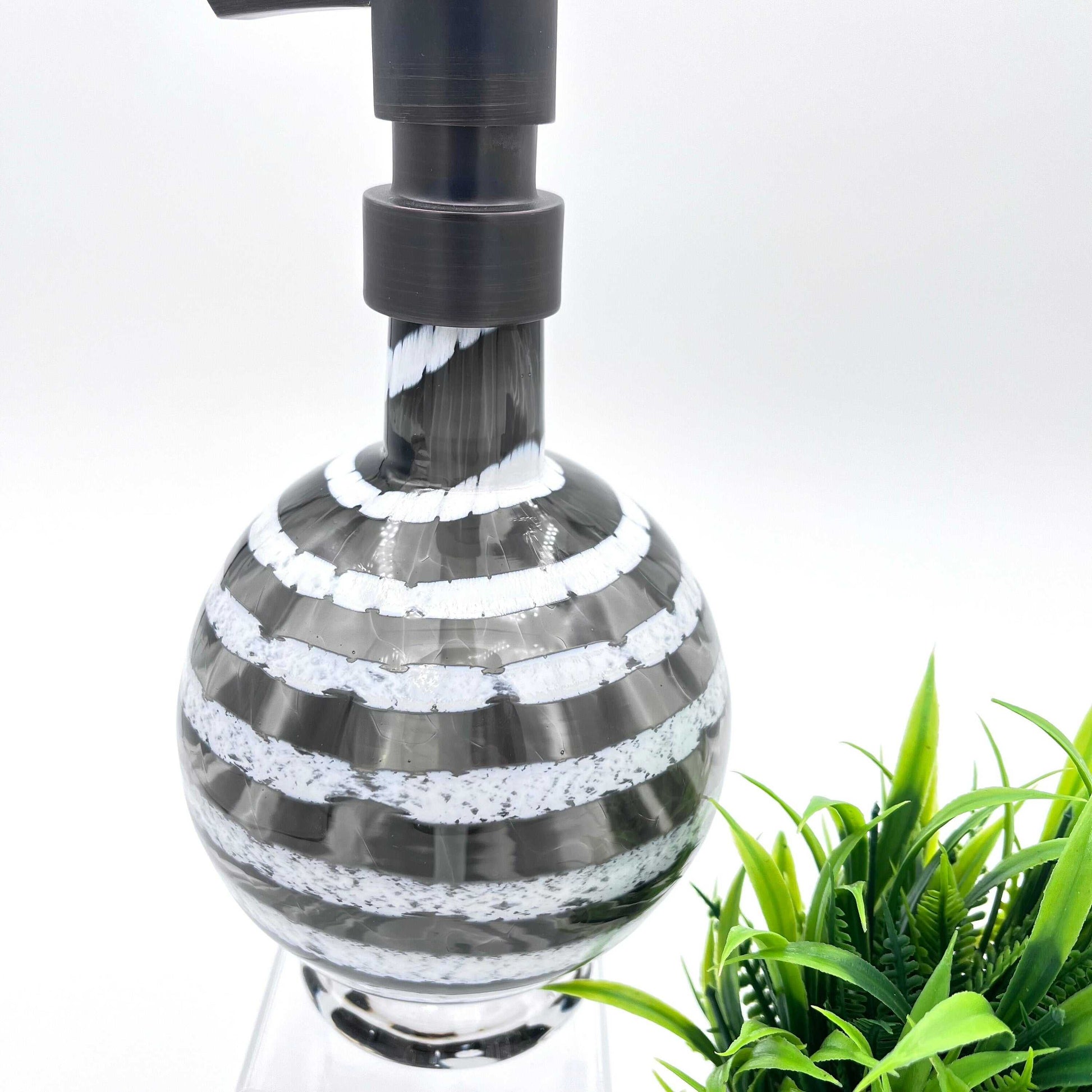 Boho handblown glass soap dispenser in gray with distressed white spiral design and black pump top near green plant
