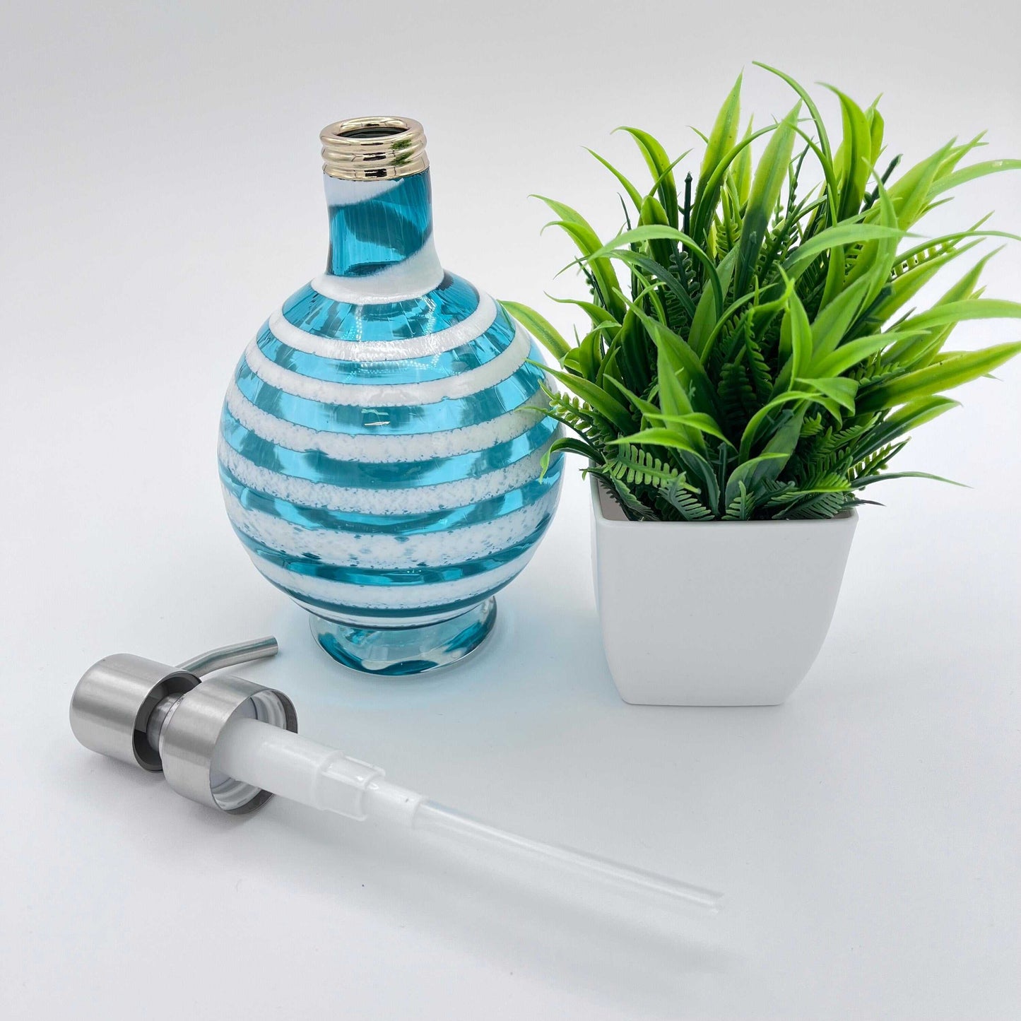 Boho handblown teal blue glass soap dispenser with distressed white spiral and removable pump next to green plant in white pot
