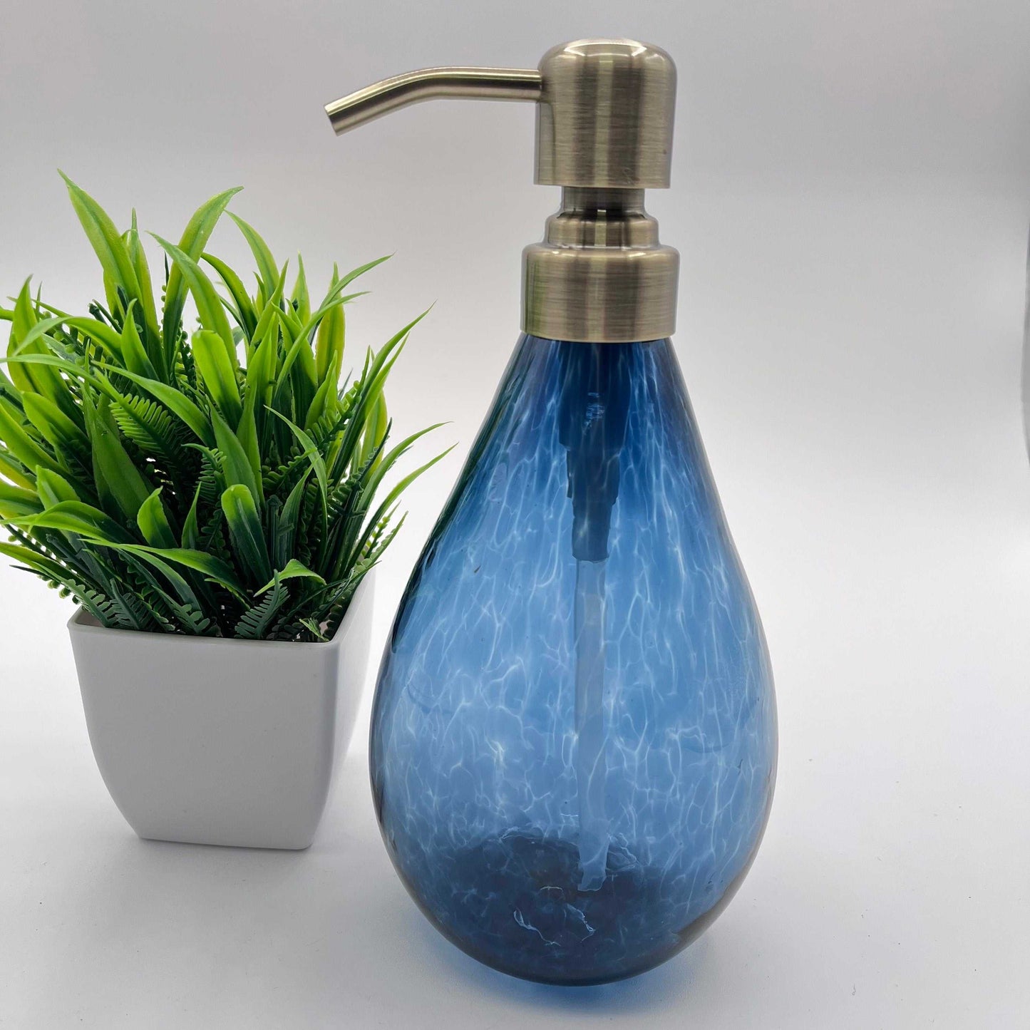 Hand blown blue glass soap dispenser with brushed metal pump and 12-ounce capacity next to small green potted plant