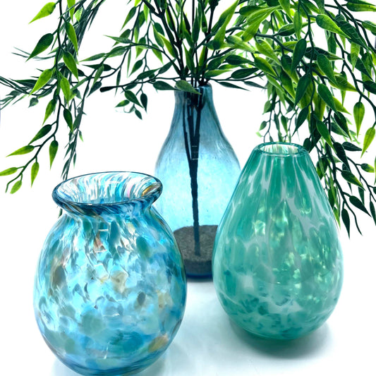 Three handmade glass vases in blue and green hues with leafy branches inside, showcasing unique textures and shapes.