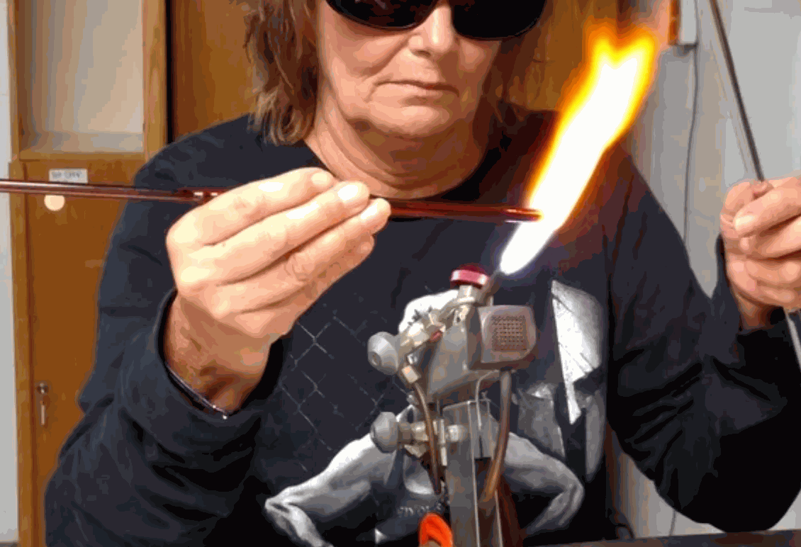 Woman wearing protective glasses shaping molten glass with a bench torch in flameworking studio during a private group glass experience