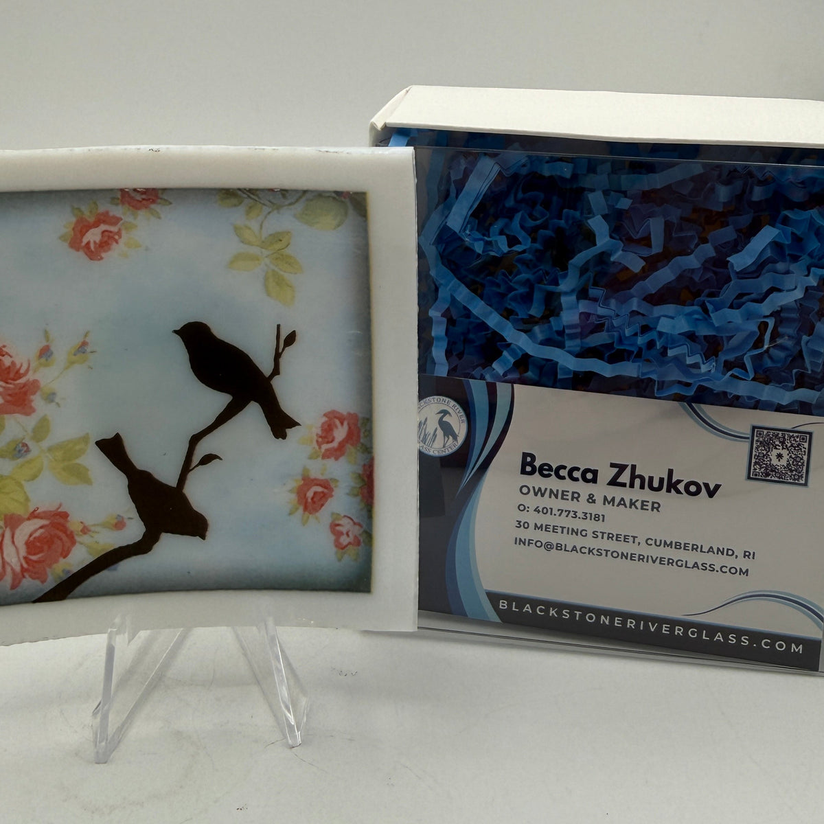 A square ceramic tile with a floral and bird design displayed on a clear stand next to its branded Becca Zhukov packaging box.