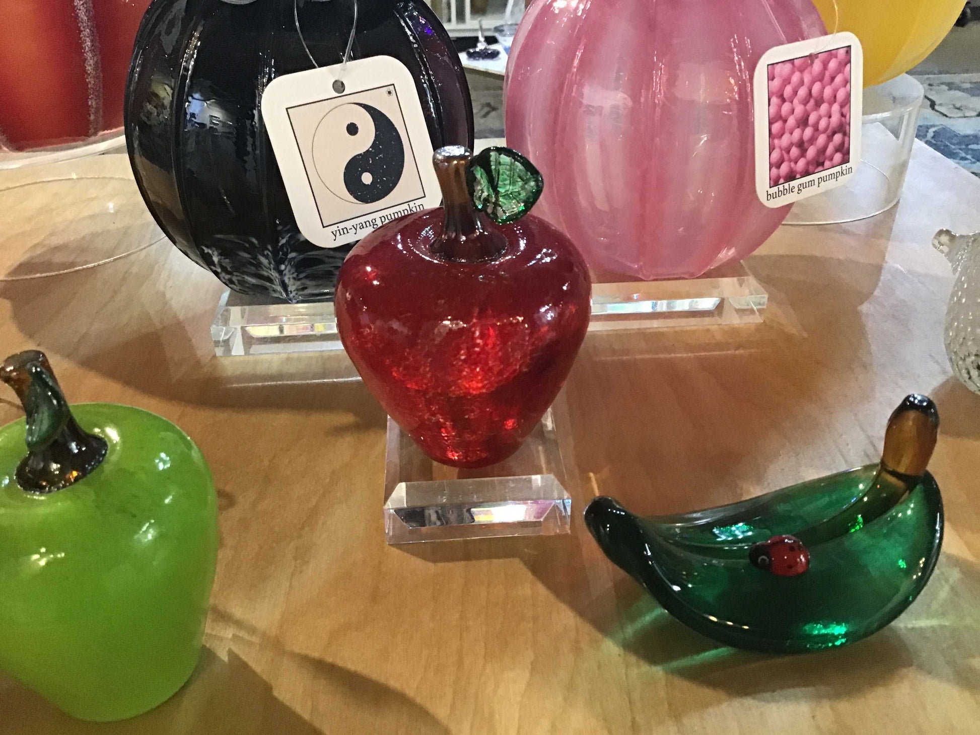 Large red apple glass ornament displayed on a wooden surface with other colorful glass fruit decorations.