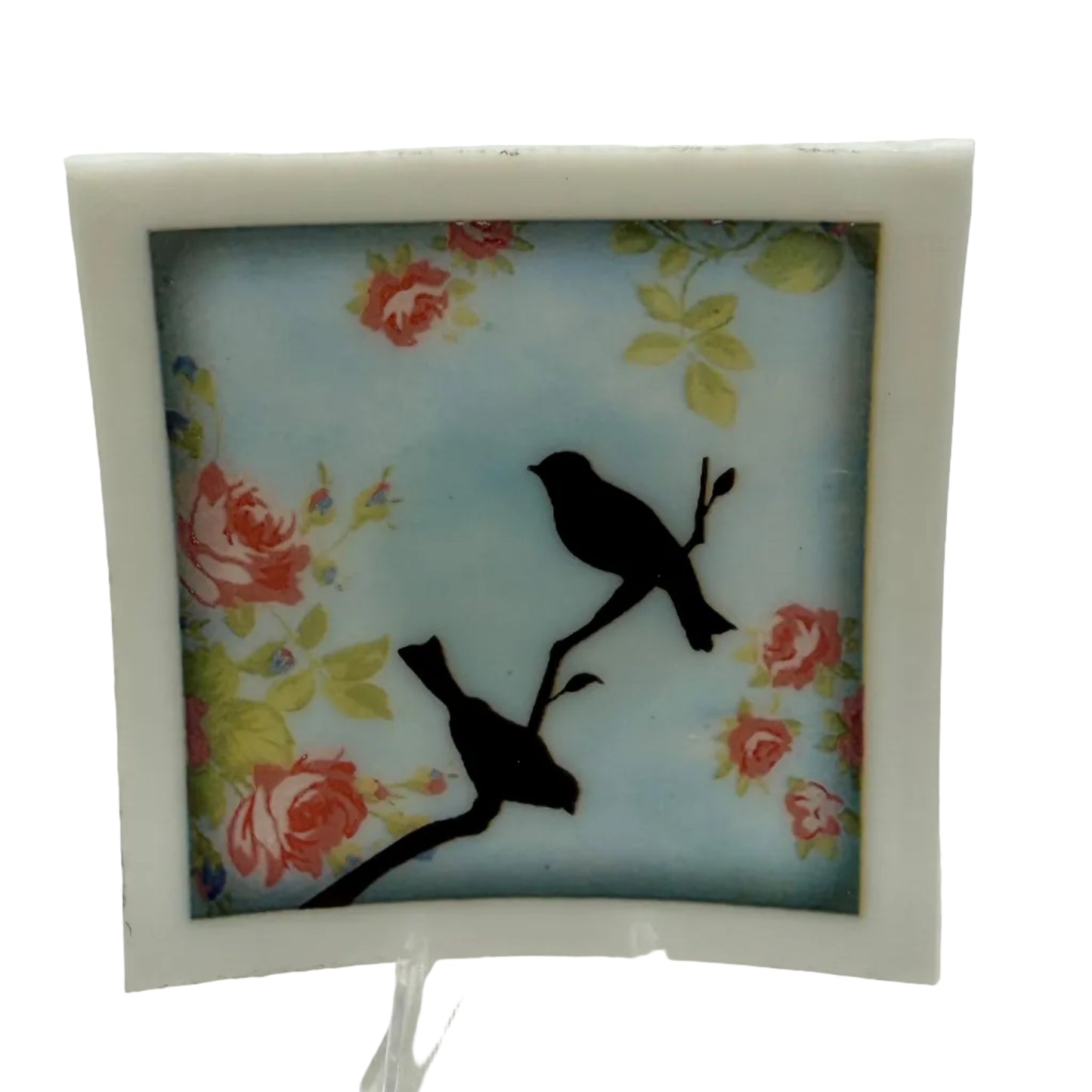 A square decorative art tile with a white border, featuring two black bird silhouettes perched on branches against a light blue background with red and yellow floral motifs, displayed on a clear stand.