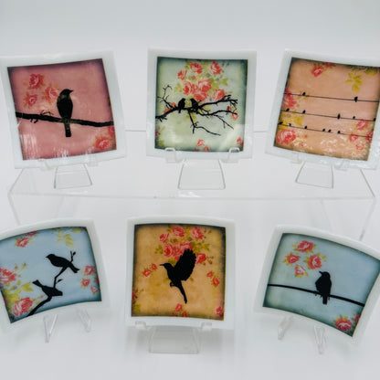 Six small square decorative glass trinket dishes with white borders are arranged on clear acrylic stands against a white background. Each tile features a different bird silhouette perched on branches or wires over floral pink and yellow backgrounds.