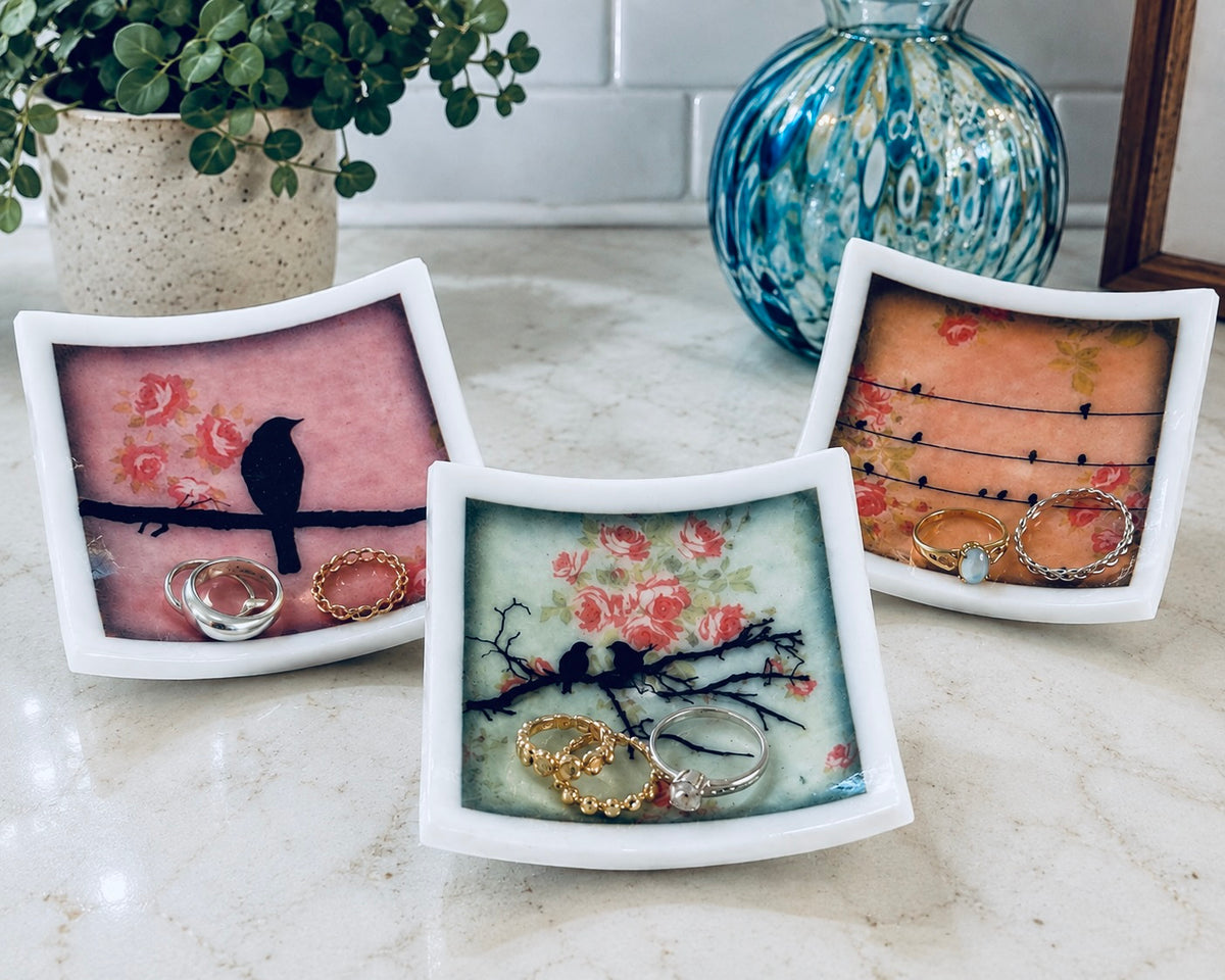 Three small square white ceramic jewelry trays with floral and bird designs, displayed on a marble countertop with rings inside them, alongside a potted plant and a blue glass vase.