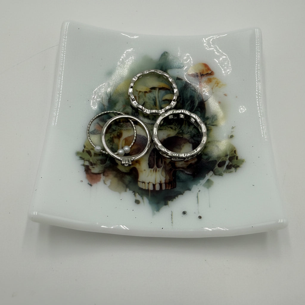 Three silver-toned rings with clear stones are displayed on a square white ceramic plate featuring a skull and floral design, set against a plain white background.