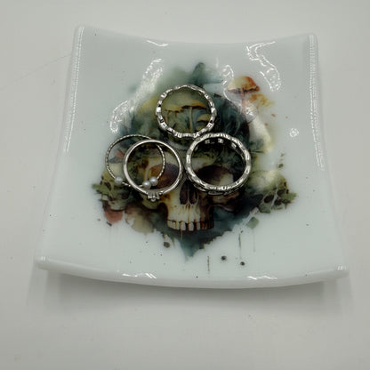 Three silver-toned rings with clear stones are displayed on a square white ceramic plate featuring a skull and floral design, set against a plain white background.