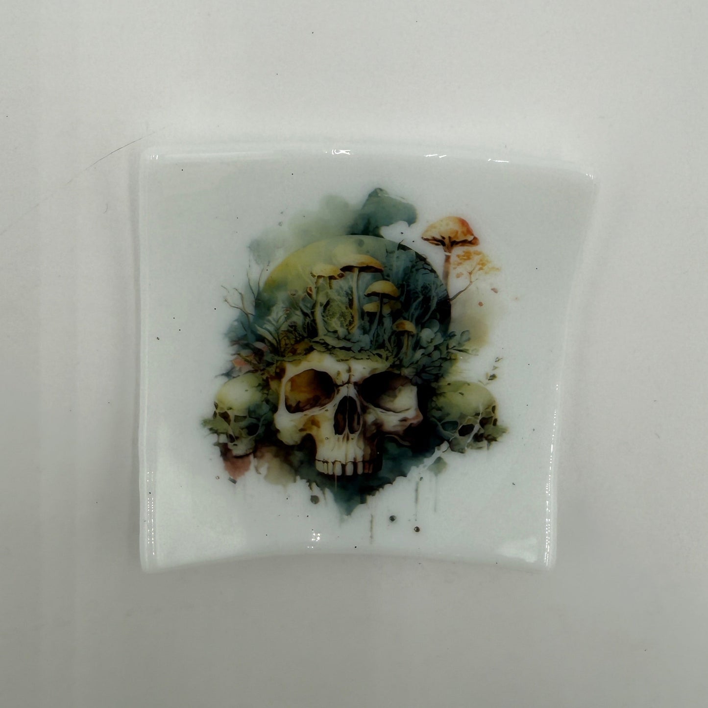 A square white glossy tile or coaster featuring a central skull illustration with a green, blue, and brown watercolor-style background, shown on a plain light gray surface.