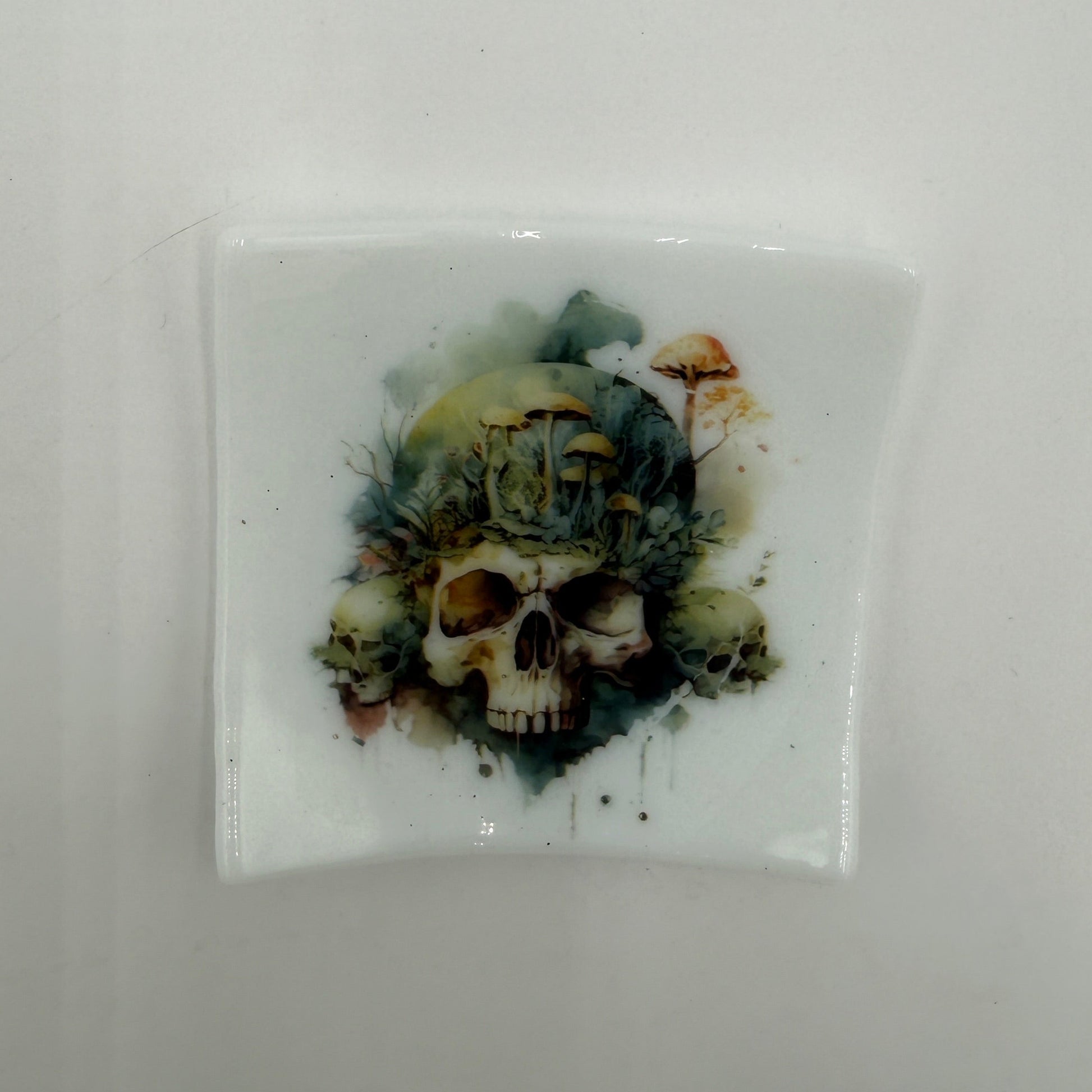 A square white glossy tile or coaster featuring a central skull illustration with a green, blue, and brown watercolor-style background, shown on a plain light gray surface.