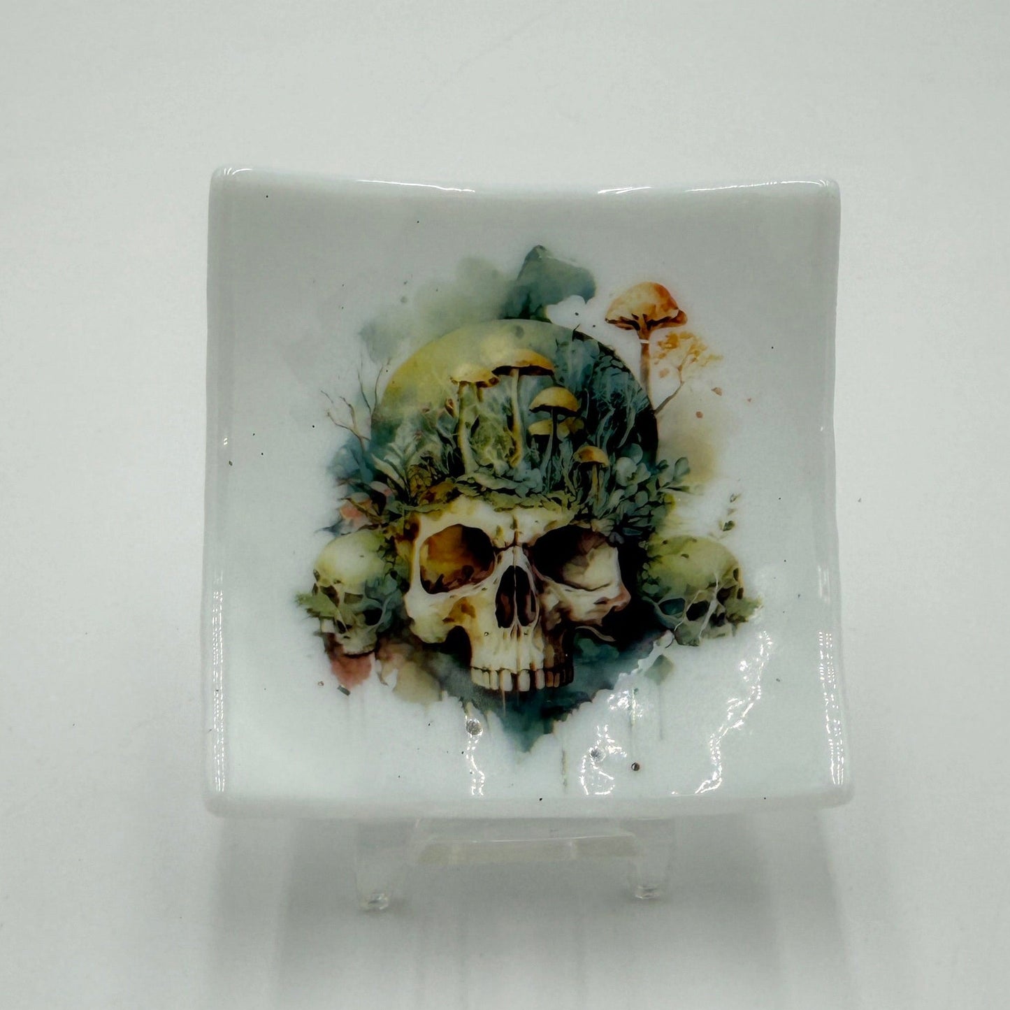 A square white glass dish featuring a detailed skull and floral design printed on the front, displayed on a small clear stand against a plain white background.