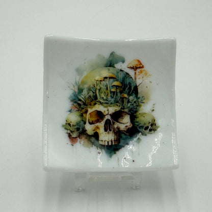 A square white glass dish featuring a detailed skull and floral design printed on the front, displayed on a small clear stand against a plain white background.