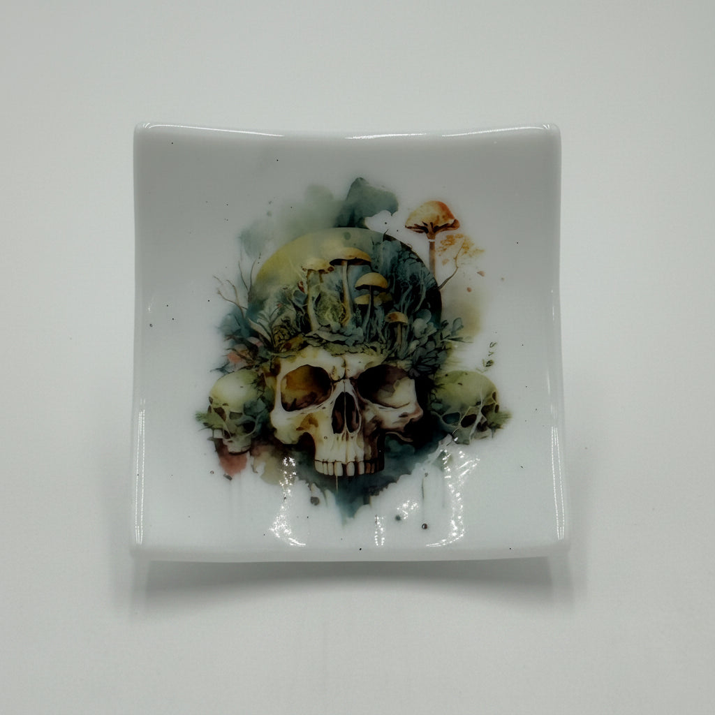 A square white ceramic dish featuring a detailed graphic of skulls and foliage in earthy green, brown, and black tones, displayed on a small clear stand against a plain white background.